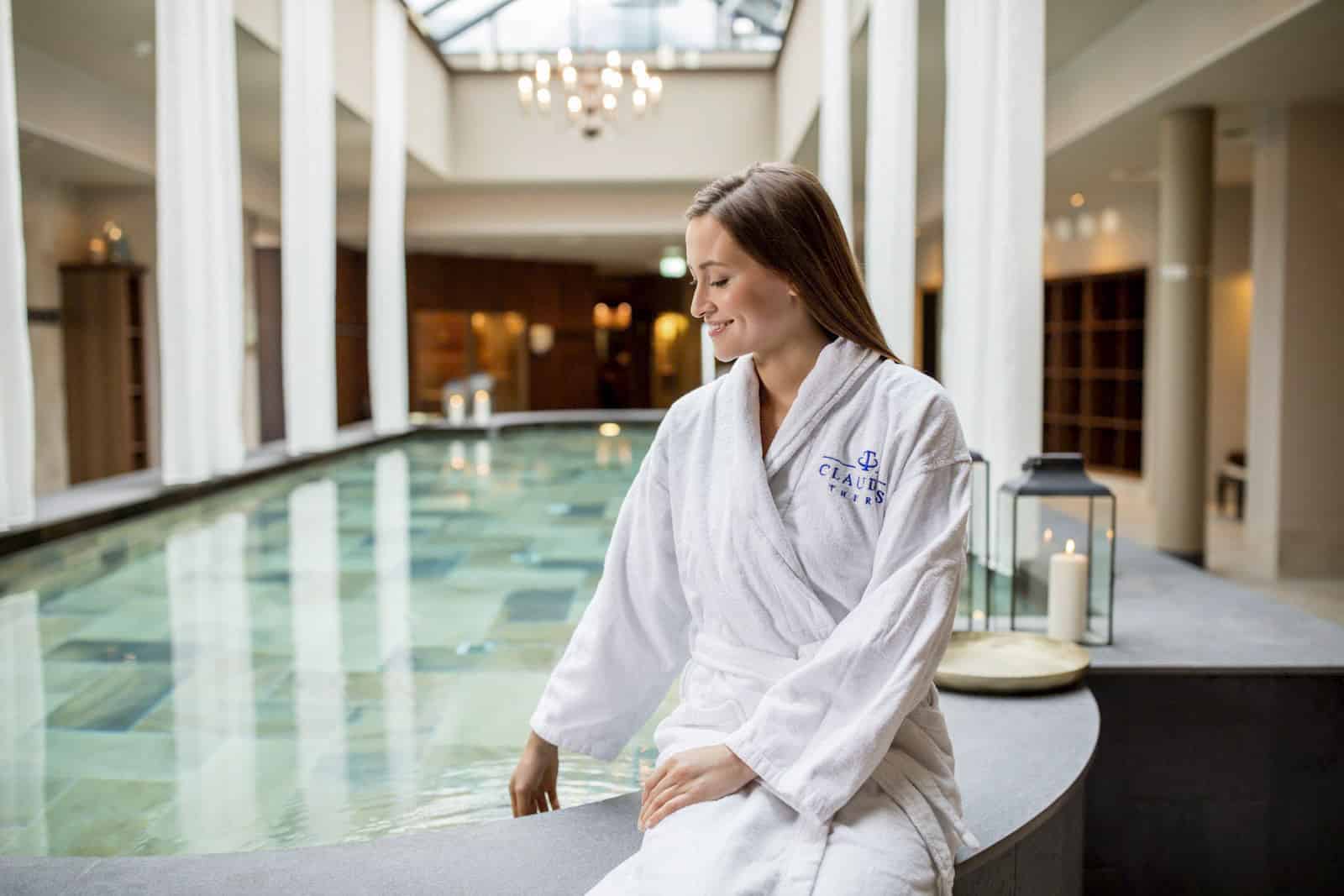 Eine Frau im weißen Bademantel sitzt am Rand eines Innenpools und berührt sanft das Wasser. Das Spa-Ambiente zeichnet sich durch hohe Fenster, sanfte Beleuchtung und Kerzen aus und schafft eine ruhige und entspannende Atmosphäre. © Fotografie Tomas Rodriguez