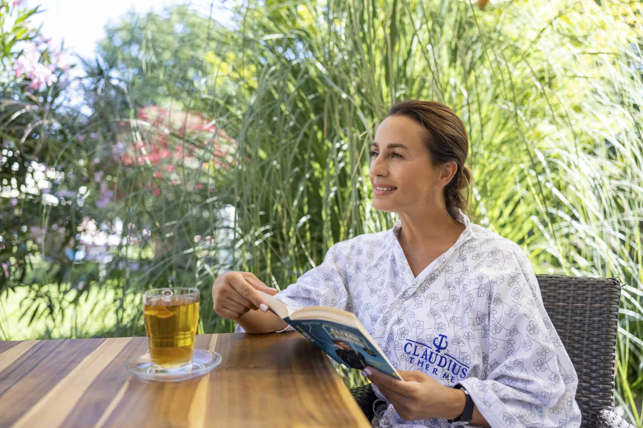 Eine Person sitzt im Freien an einem Holztisch. Sie trägt einen Bademantel mit dem Aufdruck „Claudius Therme“. Sie hält ein Buch in der Hand und lächelt. Neben ihr steht ein Glas Tee. Im Hintergrund sind hohe grüne Pflanzen zu sehen. © Fotografie Tomas Rodriguez