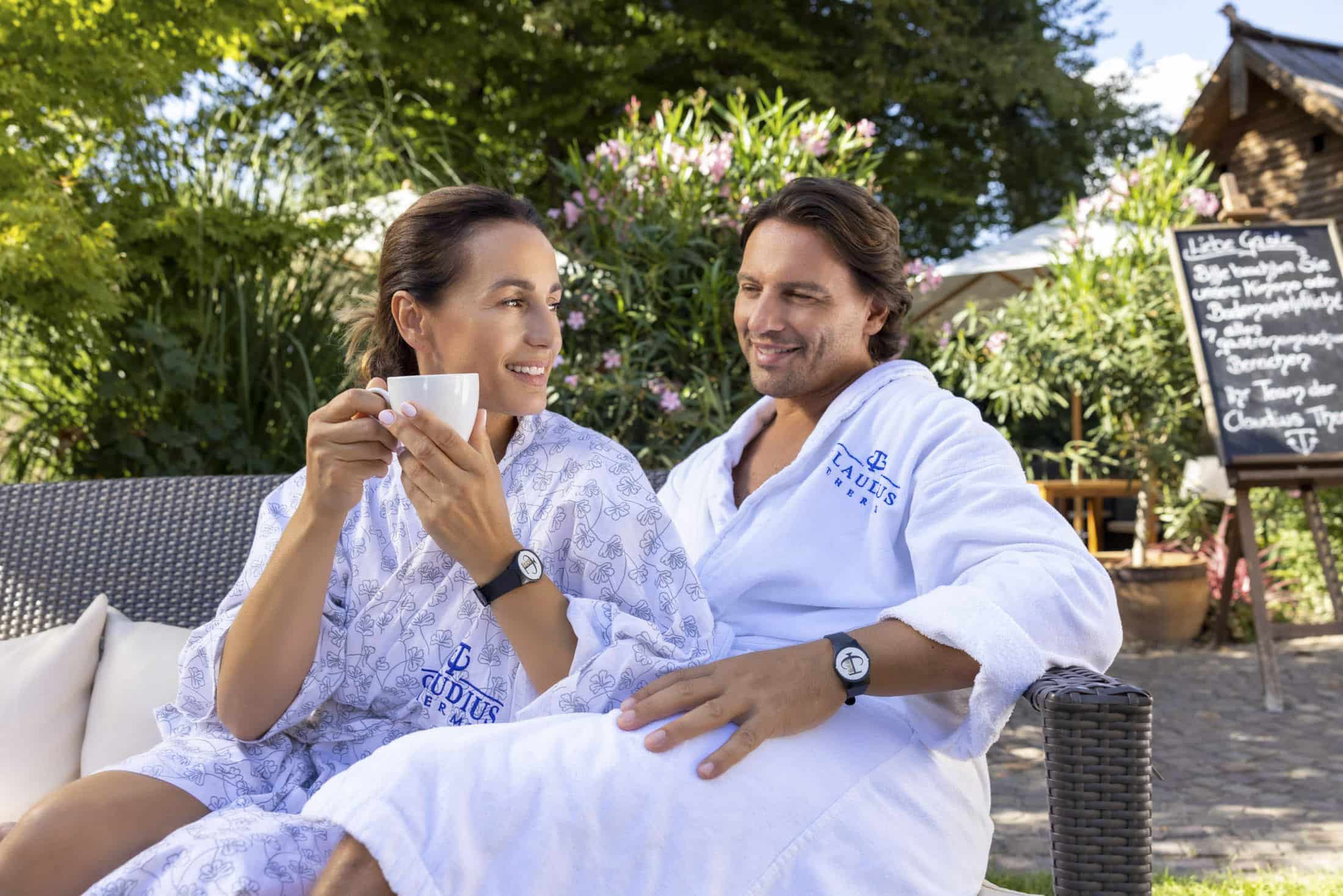 Eine Frau und ein Mann in weißen Gewändern sitzen auf Gartenmöbeln, umgeben von Grün. Die Frau hält eine Tasse und lächelt den Mann an. Im Hintergrund ist eine Tafel mit Menü zu sehen. Beide tragen passende Armbanduhren. © Fotografie Tomas Rodriguez