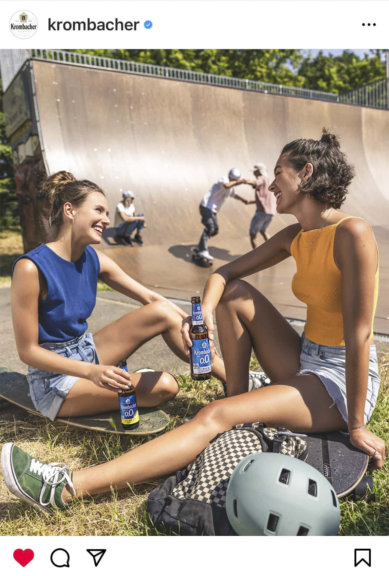 Zwei Frauen sitzen auf dem Boden neben einer Skaterampe und lächeln sich mit Krombacher-Flaschen in der Hand an. Sie tragen lässige Sommerkleidung. Ein Skateboardhelm und eine Tasche liegen auf dem Boden, im Hintergrund sind Skateboarder aktiv. © Fotografie Tomas Rodriguez