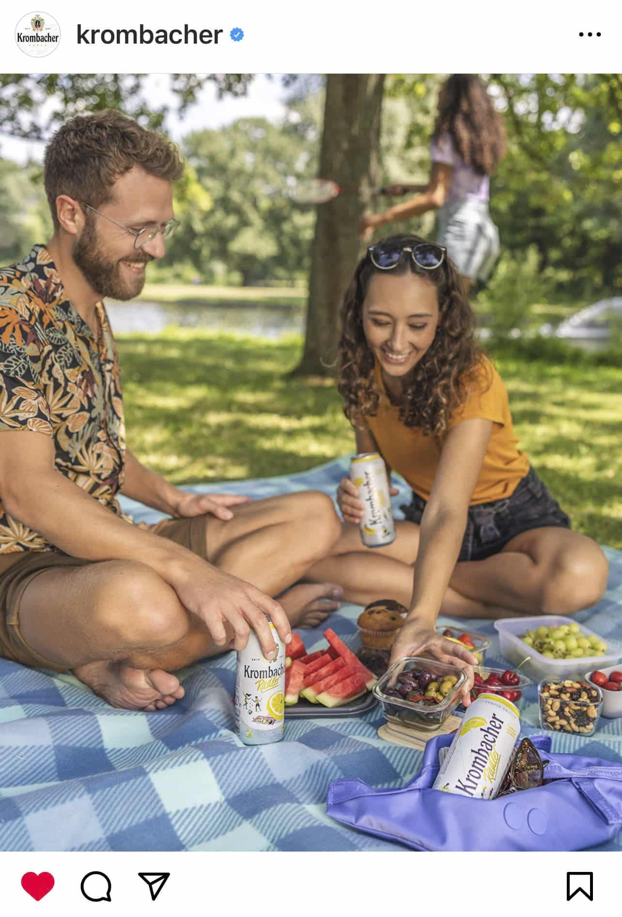 Ein Mann und eine Frau genießen ein Picknick auf einer blau karierten Decke in einem Park. Sie haben Dosen Krombacher, Wassermelonenscheiben, Trauben und andere Snacks dabei. Beide lächeln, im Hintergrund sind Bäume und Gras zu sehen. In der Ferne ist eine weitere Person zu sehen. © Fotografie Tomas Rodriguez