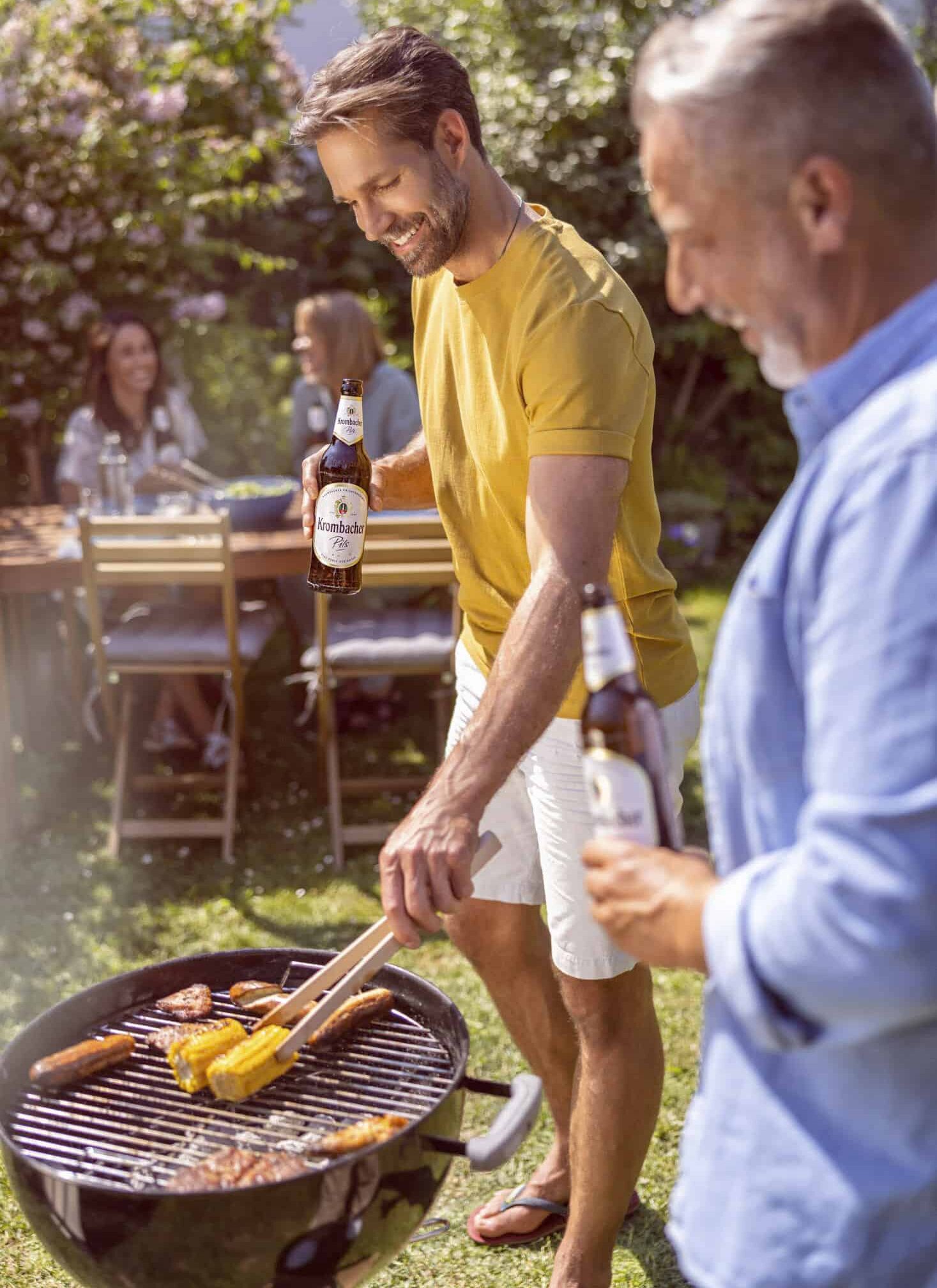 Zwei Männer grillen Würstchen und Mais auf einem Grill im Hinterhof und halten Bierflaschen in den Händen, während im Hintergrund zwei Frauen an einem Tisch sitzen. Die Szene spielt an einem sonnigen Tag mit viel Grün im Hintergrund. © Fotografie Tomas Rodriguez