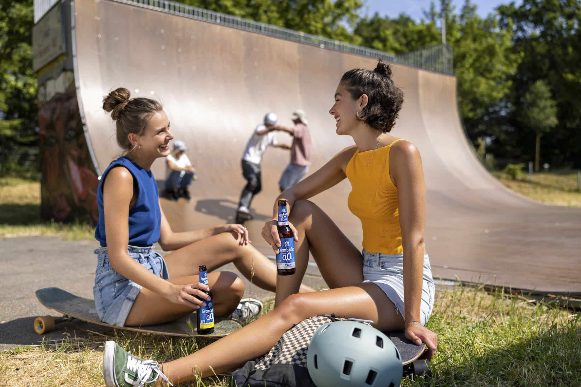 Zwei Frauen in lässiger Sommerkleidung sitzen auf dem Rasen neben einer Skateboard-Rampe und halten jeweils eine Flasche in der Hand. Sie lächeln und unterhalten sich. Im Hintergrund üben Skateboarder auf der Rampe. In der Nähe liegt ein Helm. © Fotografie Tomas Rodriguez