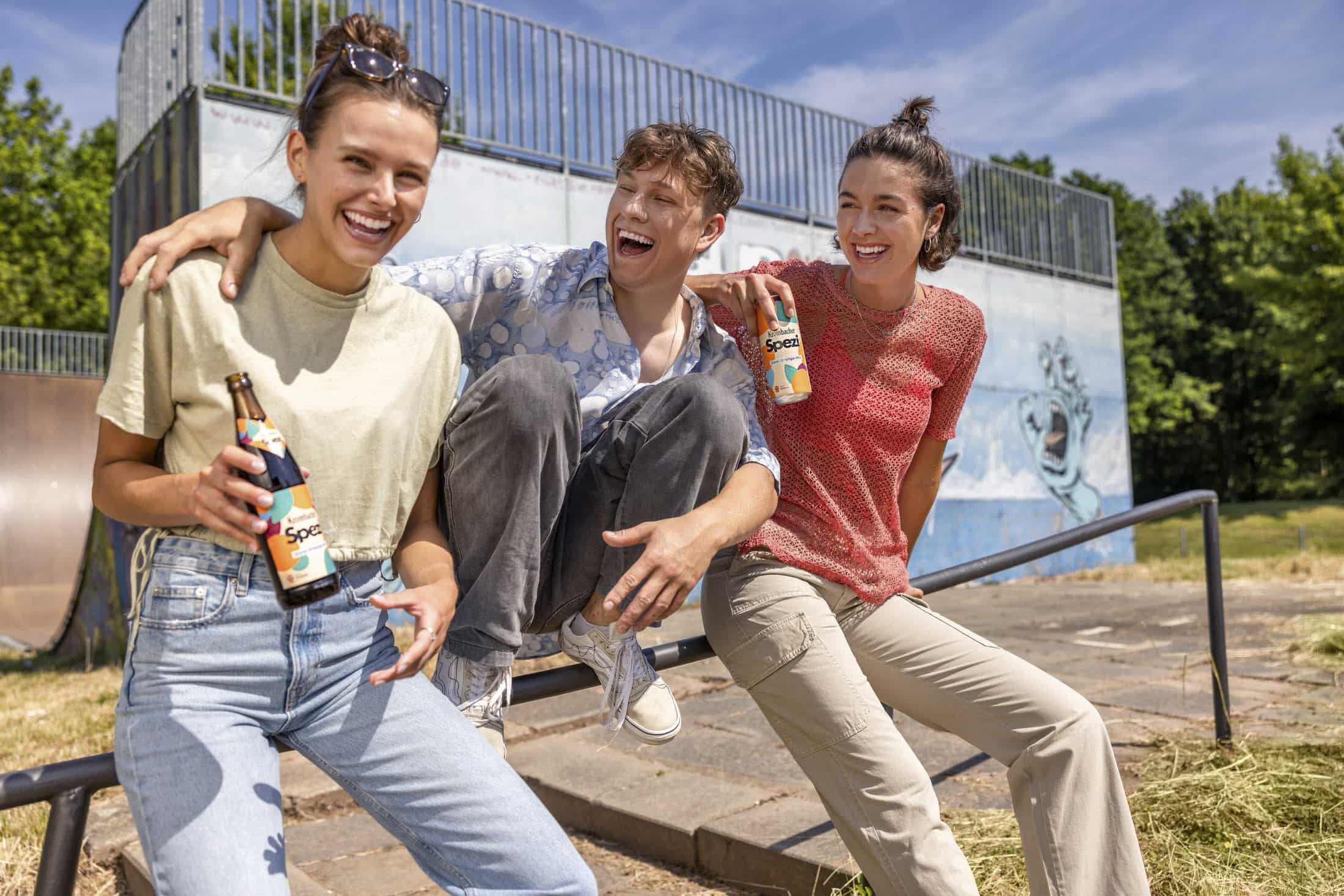 Drei Freunde genießen Drinks in einem Skatepark. Die Person in der Mitte sitzt halb auf einem Geländer, während die anderen neben ihnen stehen. Sie sind im Freien, mit einer mit Graffiti bedeckten Rampe und grünen Bäumen im Hintergrund. © Fotografie Tomas Rodriguez
