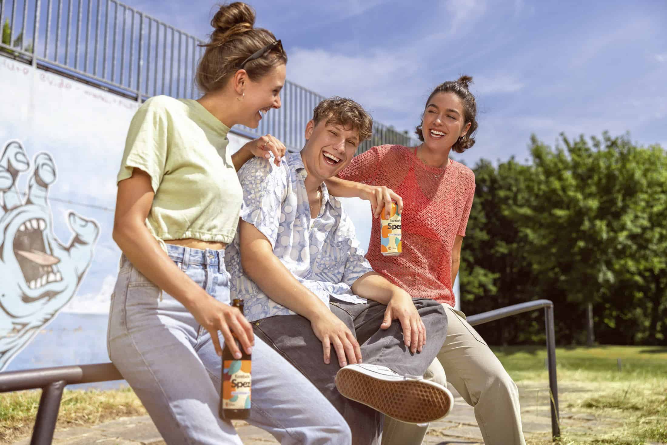 Drei Menschen sitzen im Freien zusammen, lachen und halten Getränkeflaschen in den Händen. Zwei von ihnen sind Frauen, die helle Oberteile und Jeans tragen, während der Mann in der Mitte ein gemustertes Hemd trägt. Im Hintergrund ist ein Wandgemälde mit einer Hand auf einer blauen Wand zu sehen. © Fotografie Tomas Rodriguez