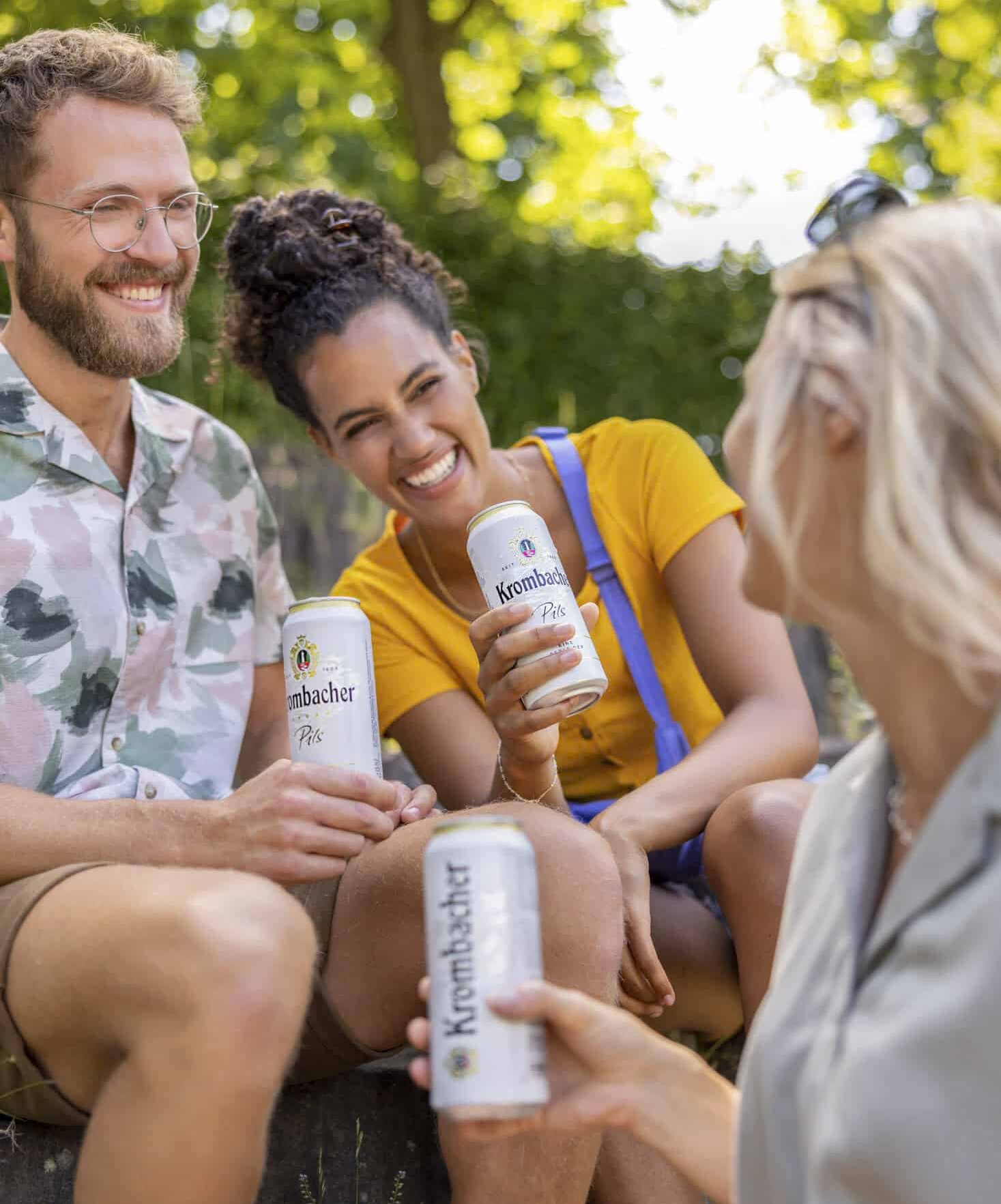Drei Freunde sitzen an einem sonnigen Tag im Freien und halten alle Dosen Krombacher in der Hand. Eine Frau mit Brille blickt weg, eine andere Frau in Gelb lacht und ein Mann mit Bart lächelt. Im Hintergrund sind Bäume und Grün zu sehen. © Fotografie Tomas Rodriguez