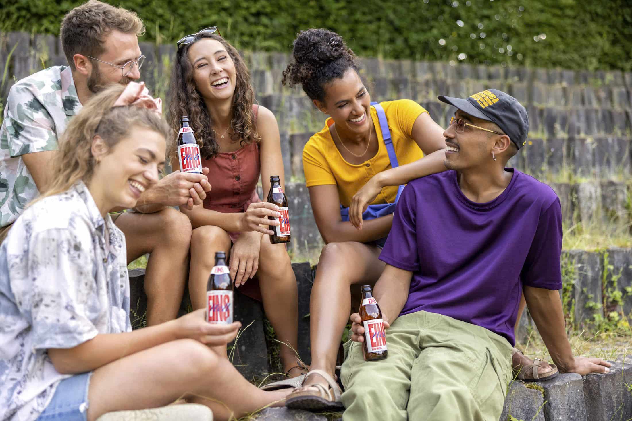 Eine Gruppe von fünf Personen sitzt draußen auf Steinstufen und genießt Getränke aus der Flasche. Sie lachen und lächeln und suggerieren damit eine entspannte, fröhliche Atmosphäre. Der Hintergrund ist grün mit Laub. © Fotografie Tomas Rodriguez
