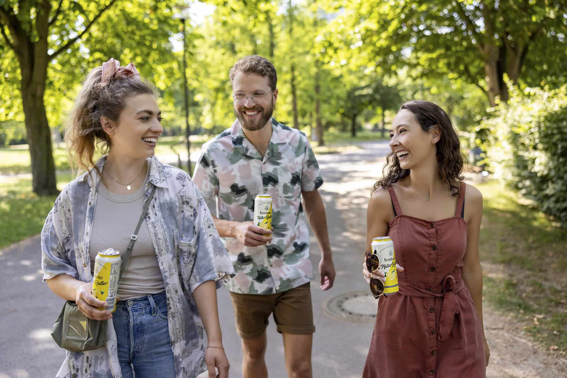 Drei Personen gehen an einem sonnigen Tag im Freien spazieren und halten Getränkedosen in der Hand. Die Person links trägt ein gemustertes Hemd und Jeans, die Person in der Mitte trägt ein geblümtes Hemd und Shorts und die Person rechts trägt ein ärmelloses Kleid. Sie lächeln alle. © Fotografie Tomas Rodriguez