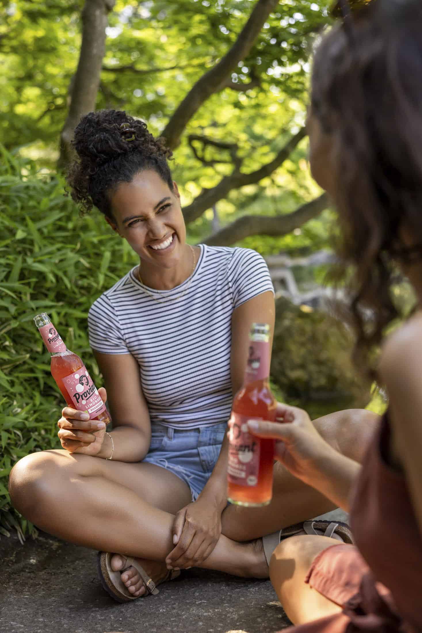 Zwei Personen sitzen an einem sonnigen Tag im Freien und halten rosa Getränkeflaschen in den Händen. Eine Person trägt ein gestreiftes Hemd und Jeansshorts und lächelt, während sie im Schneidersitz sitzt. Im Hintergrund sind grüne Blätter zu sehen. © Fotografie Tomas Rodriguez