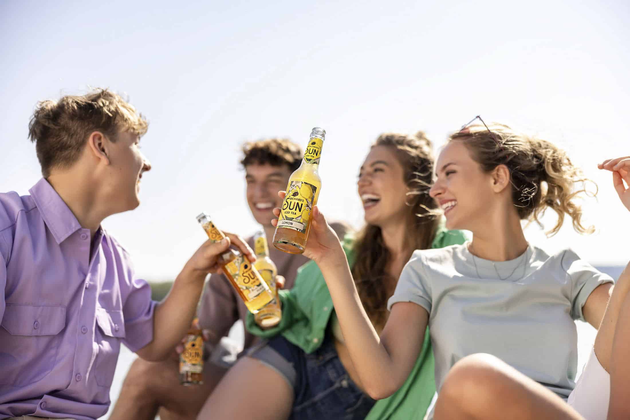 Vier Menschen lächeln und genießen im Freien Flaschengetränke, im Hintergrund ein heller Himmel. Sie wirken entspannt, tragen lässige Sommerkleidung und ihre fröhlichen Ausdrücke deuten auf eine lustige, gesellige Atmosphäre hin. © Fotografie Tomas Rodriguez