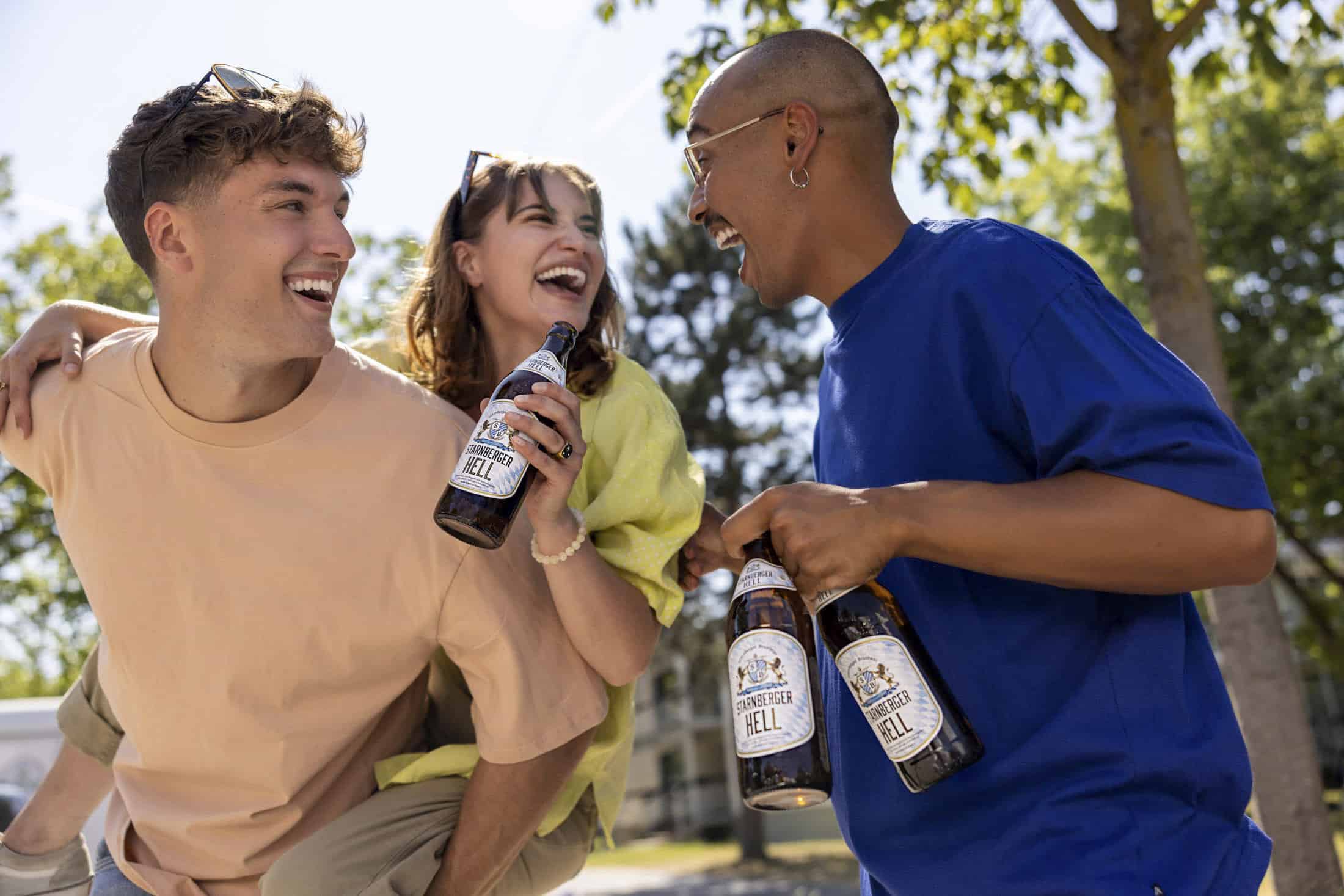 Drei Freunde lachen und trinken Bier in einem Park. Einer sitzt huckepack auf dem anderen und hält eine Flasche in der Hand. Der dritte lächelt und hält zwei Flaschen in der Hand. Sie sind von Bäumen und Sonnenlicht umgeben. © Fotografie Tomas Rodriguez