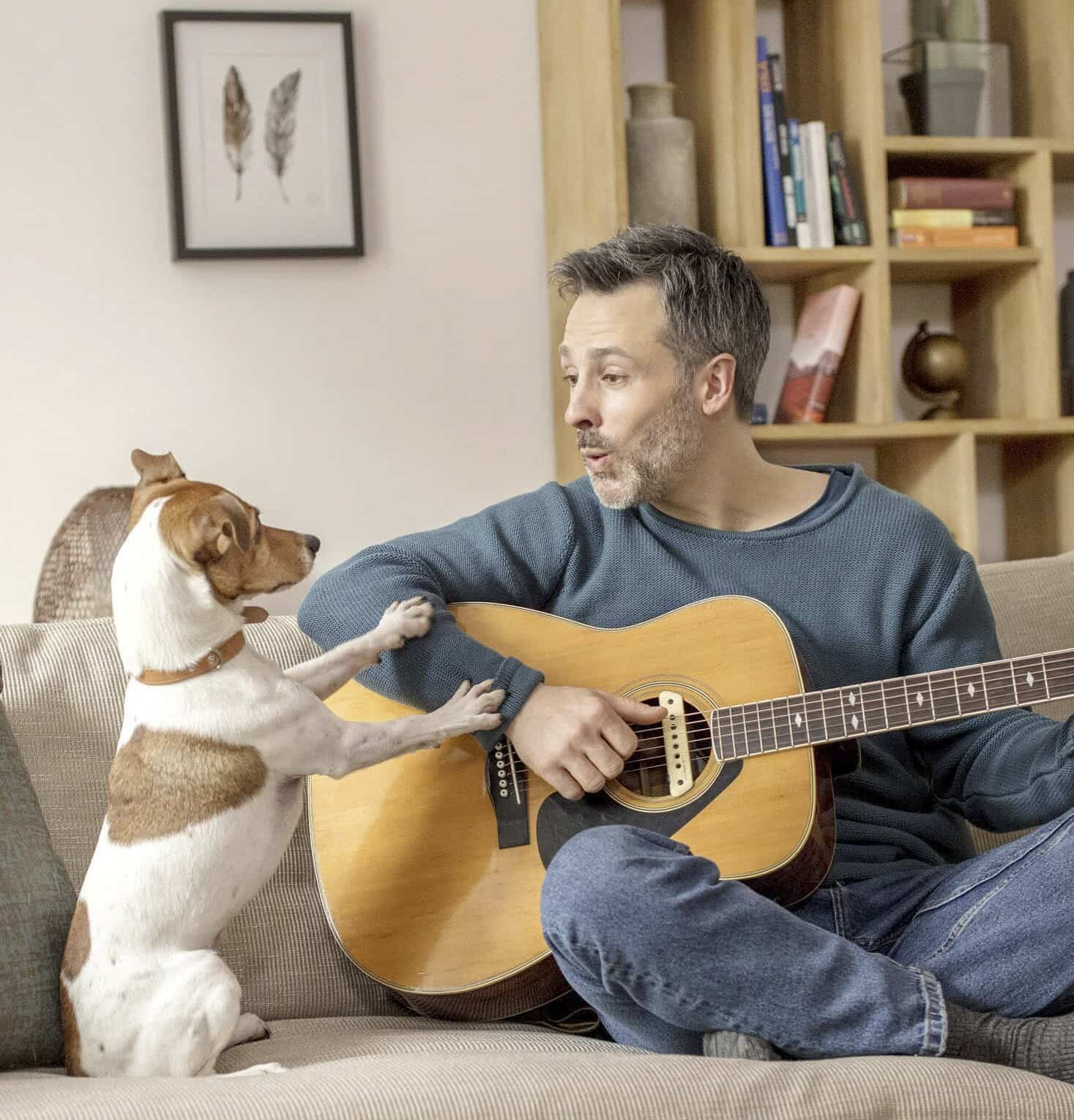 Ein Mann mit grauem Haar und Bart sitzt auf einem Sofa und hält eine Gitarre in der Hand. Neben ihm steht ein kleiner braun-weißer Hund auf seinen Hinterbeinen, die Vorderpfoten stützen sich auf sein Bein. Im Hintergrund sind ein Bücherregal und ein gerahmtes Kunstwerk zu sehen. © Fotografie Tomas Rodriguez