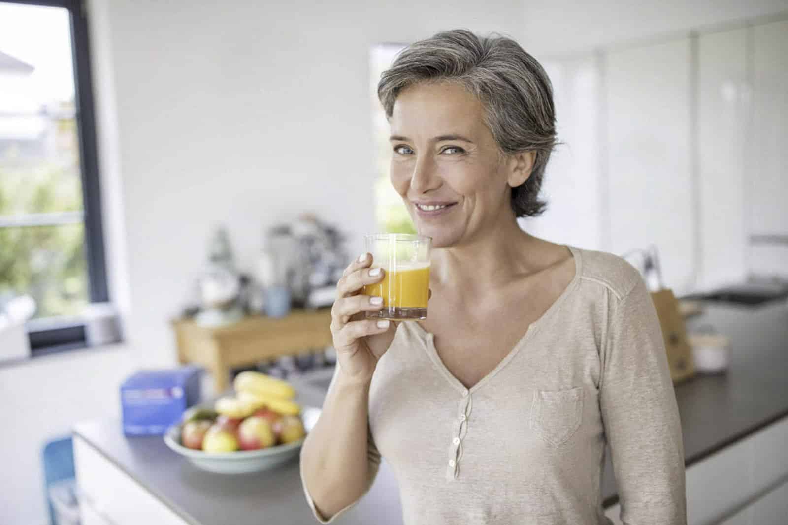 Eine Frau mit kurzen grauen Haaren lächelt und trinkt ein Glas Orangensaft in einer Küche. Auf der Theke hinter ihr steht eine Schale mit verschiedenen Früchten. Sie trägt ein hellbeiges Langarmshirt. © Fotografie Tomas Rodriguez