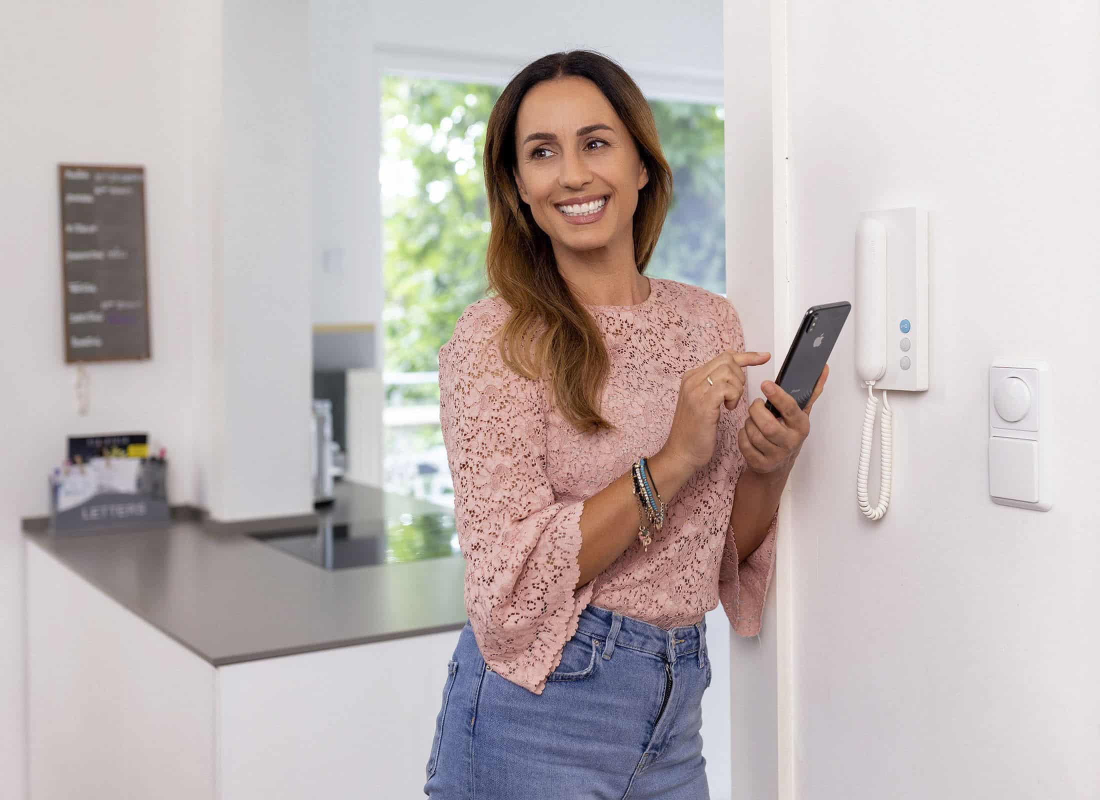 Eine Frau mit langen braunen Haaren, einer rosa Spitzenbluse und Jeans trägt, lächelt und benutzt ein Smartphone in einer modernen Küche. Sie steht neben einer Gegensprechanlage an der Wand, im Hintergrund sind eine Arbeitsplatte und ein Fenster zu sehen. © Fotografie Tomas Rodriguez