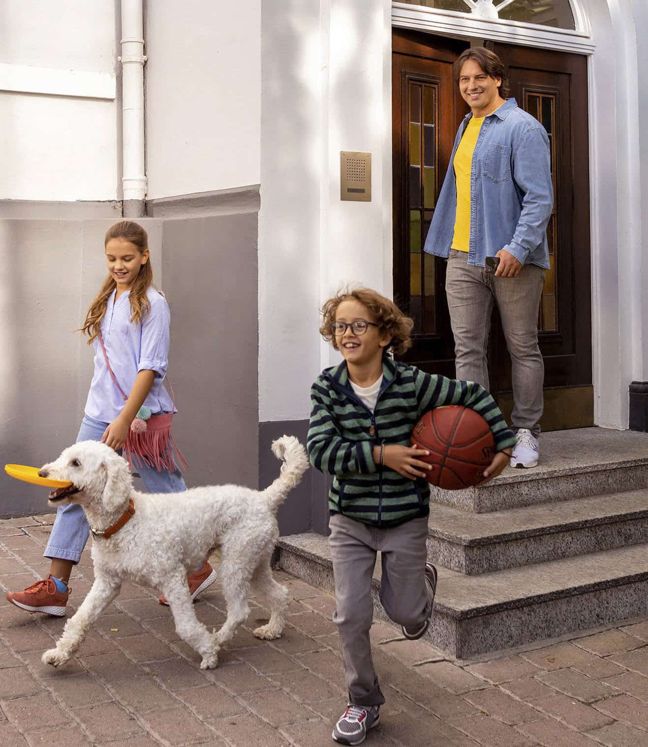 Ein Mann steht lächelnd an der Tür, während ein Mädchen mit einer Fransentasche und einem Hund mit einem Frisbee vorbeigeht. Ein Junge mit Brille läuft voraus und hält einen Basketball. Sie stehen auf einem gepflasterten Weg vor einem Gebäude. © Fotografie Tomas Rodriguez
