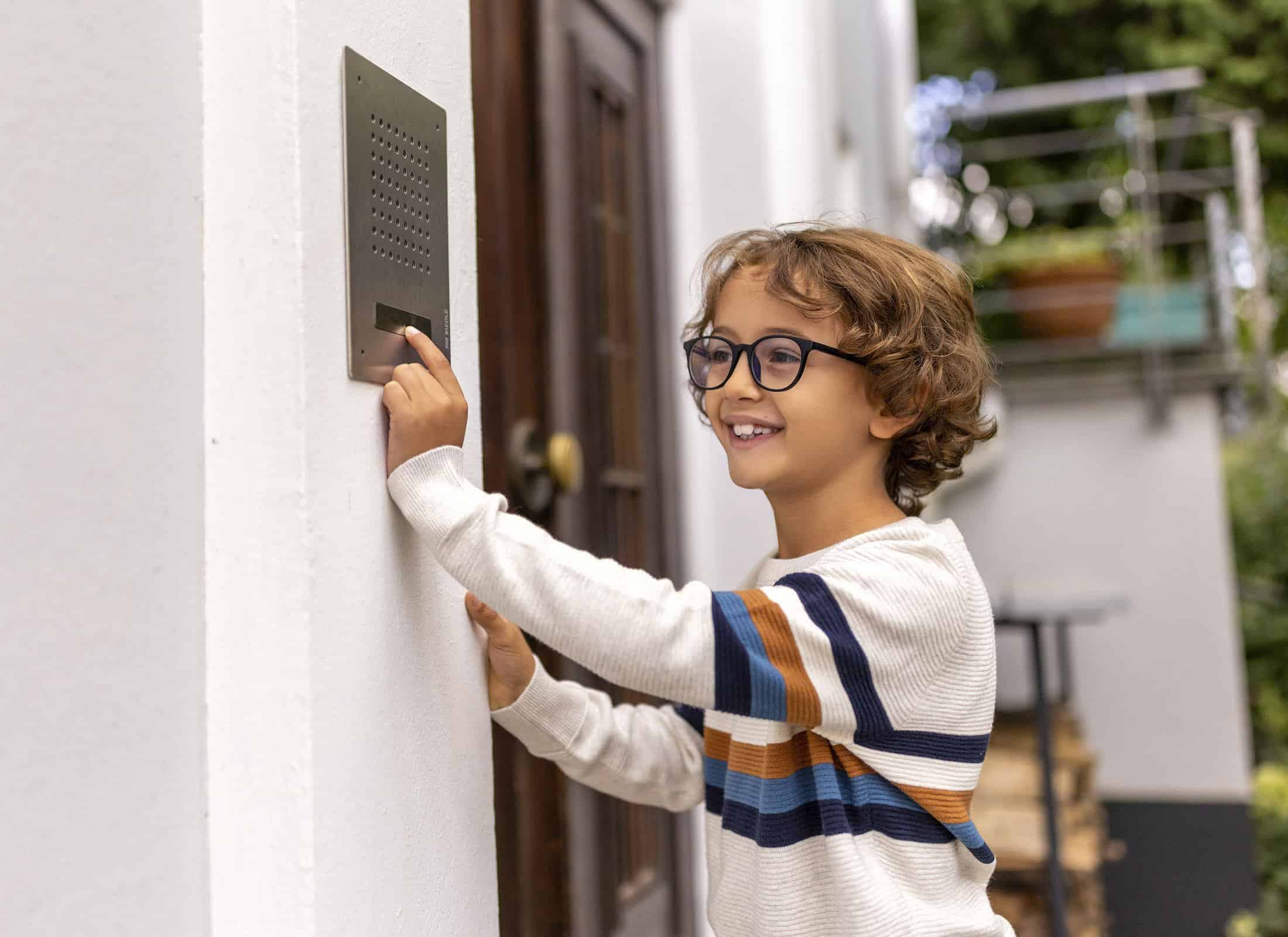 Ein lächelndes Kind mit lockigem Haar und Brille drückt einen Knopf an einer Gegensprechanlage, die an einer weißen Wand außerhalb eines Gebäudes angebracht ist. Das Kind trägt einen weißen Pullover mit blauen und orangefarbenen Streifen. Im Hintergrund sind eine Tür und verschwommenes Grün zu sehen. © Fotografie Tomas Rodriguez