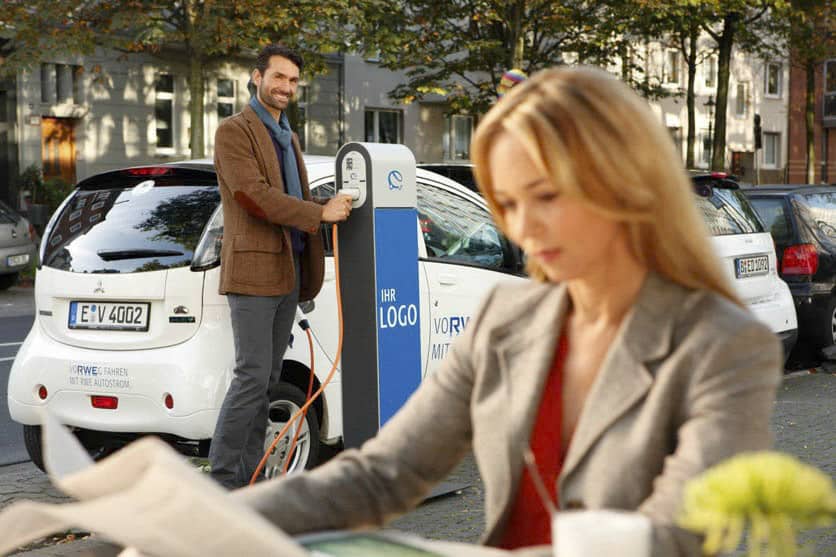Eine Person lädt ein Elektroauto an einer öffentlichen Ladestation in einer von Bäumen gesäumten Straße auf. Das Auto hat ein sichtbares Nummernschild. Eine weitere Person im Vordergrund sitzt an einem Tisch und liest eine Zeitung; der Fokus ist unscharf. © Fotografie Tomas Rodriguez