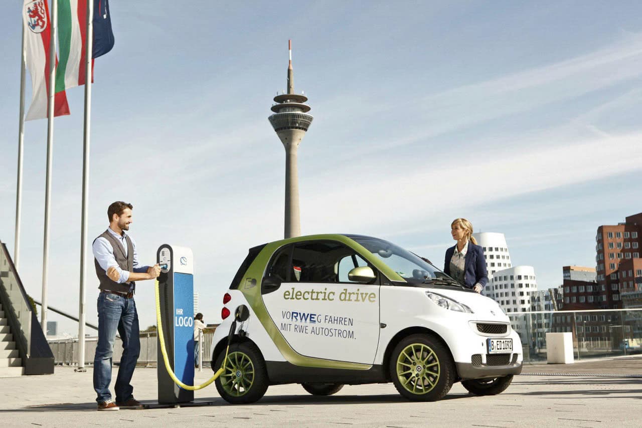 Ein Mann und eine Frau laden an einer Station in einer städtischen Umgebung ein kleines Elektroauto auf. Das Auto ist mit dem Schriftzug „Electric Drive“ versehen. Im Hintergrund sind hohe moderne Gebäude und ein markanter Turm zu sehen, in der Nähe hängen Flaggen an Masten. © Fotografie Tomas Rodriguez