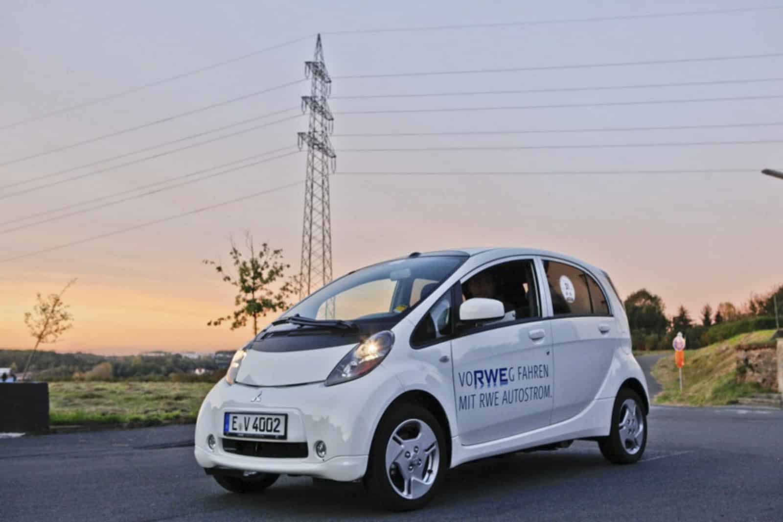 Ein kleines weißes Elektroauto parkt bei Sonnenuntergang auf einer Straße. Auf der Seite des Autos steht der Text „VORWEG FAHREN MIT RWE AUTOSTROM“. Im Hintergrund sind Stromleitungen und Bäume zu sehen. © Fotografie Tomas Rodriguez