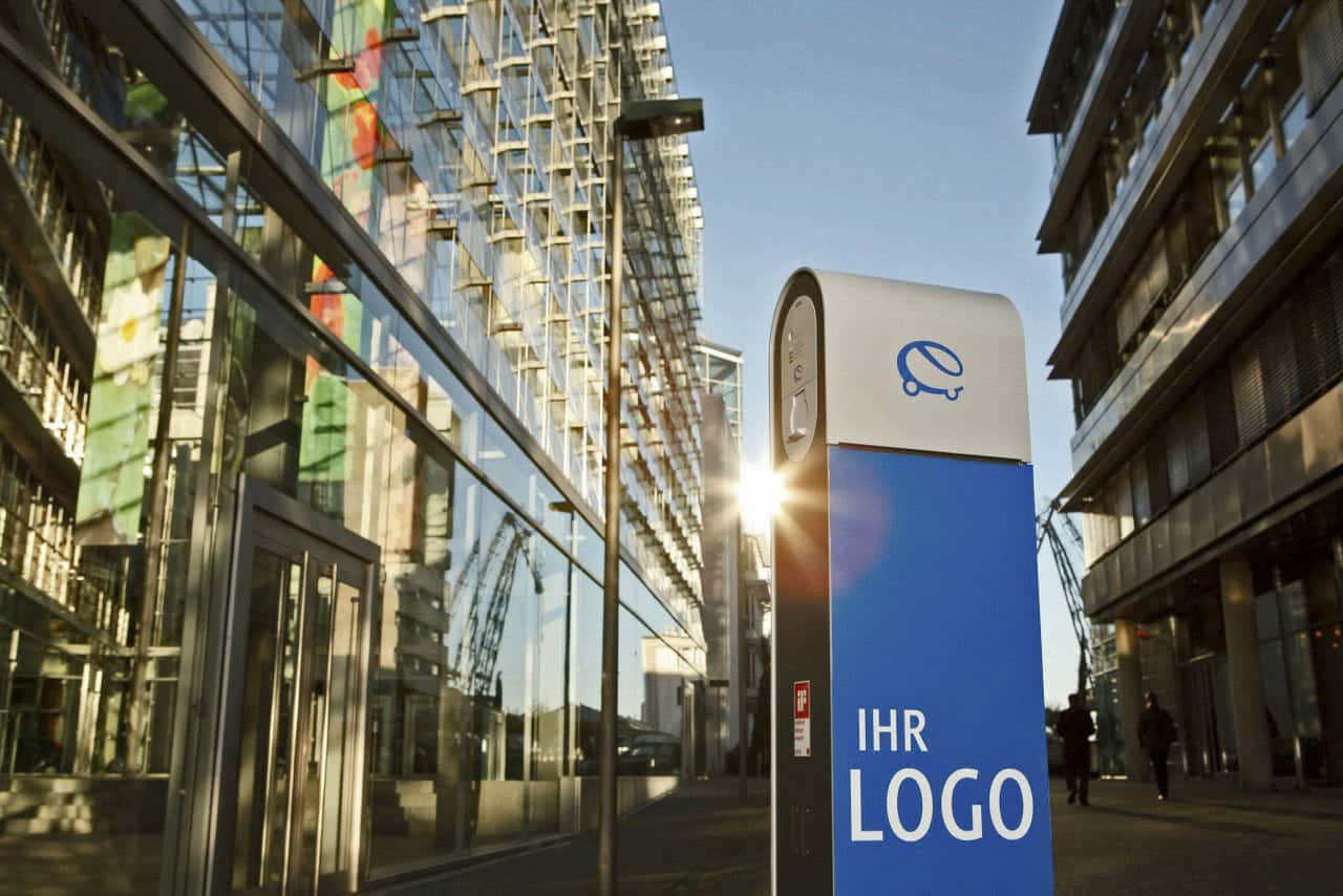 Eine moderne Stadtlandschaft mit Glasgebäuden reflektiert das Sonnenlicht. Im Vordergrund zeigt ein blau-weißer Sockel ein stilisiertes „e“-Logo und den Text „IHR LOGO“. Die Sonne ist zwischen zwei Gebäuden teilweise sichtbar. © Fotografie Tomas Rodriguez