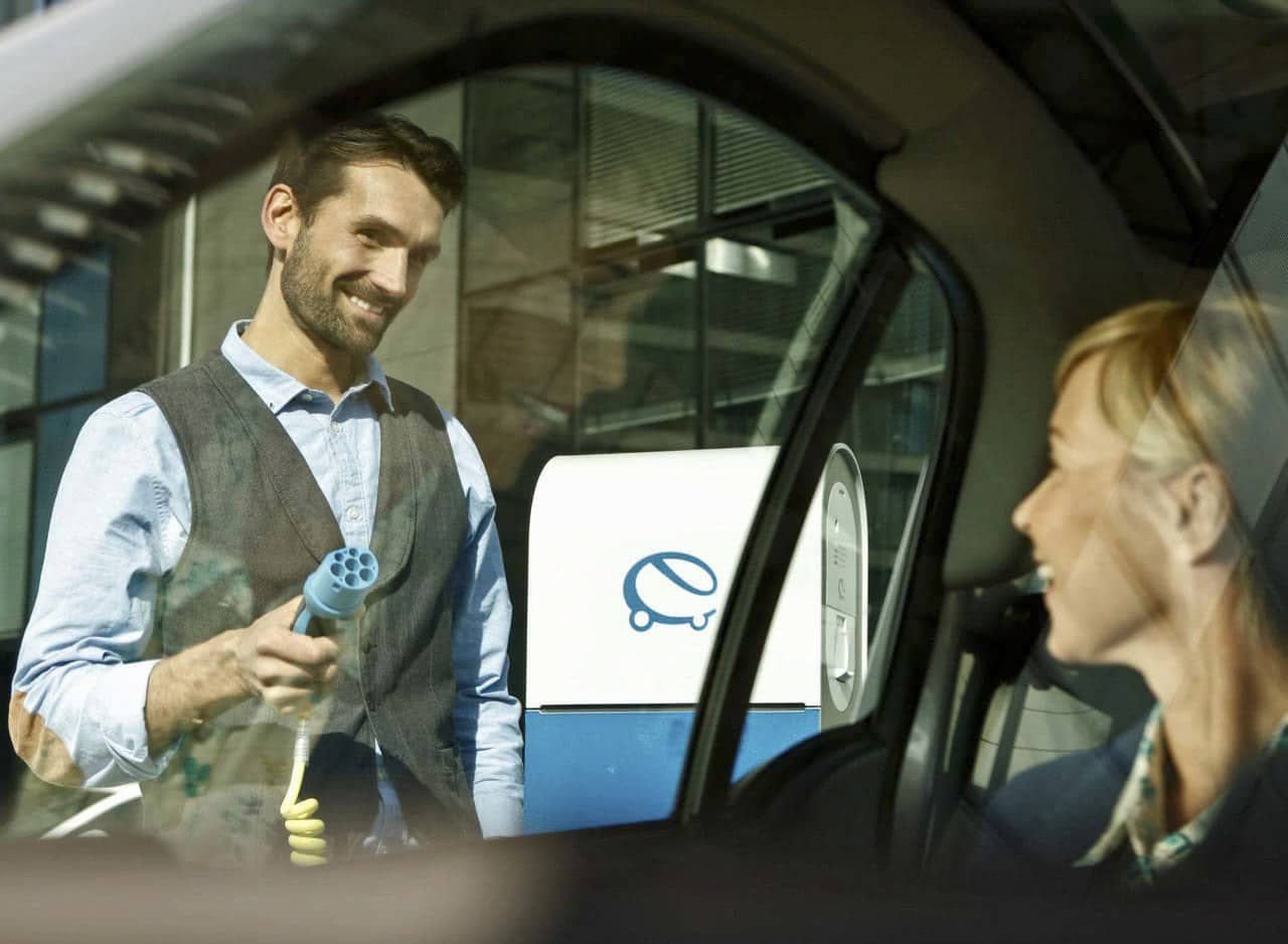 Ein Mann lächelt und hält einen Ladestecker für ein Elektroauto an einer Ladestation, sichtbar durch ein Autofenster. Eine Frau im Auto schaut ihn lächelnd an. Im Hintergrund ist ein Gebäude mit Glas- und Metallelementen zu sehen. © Fotografie Tomas Rodriguez