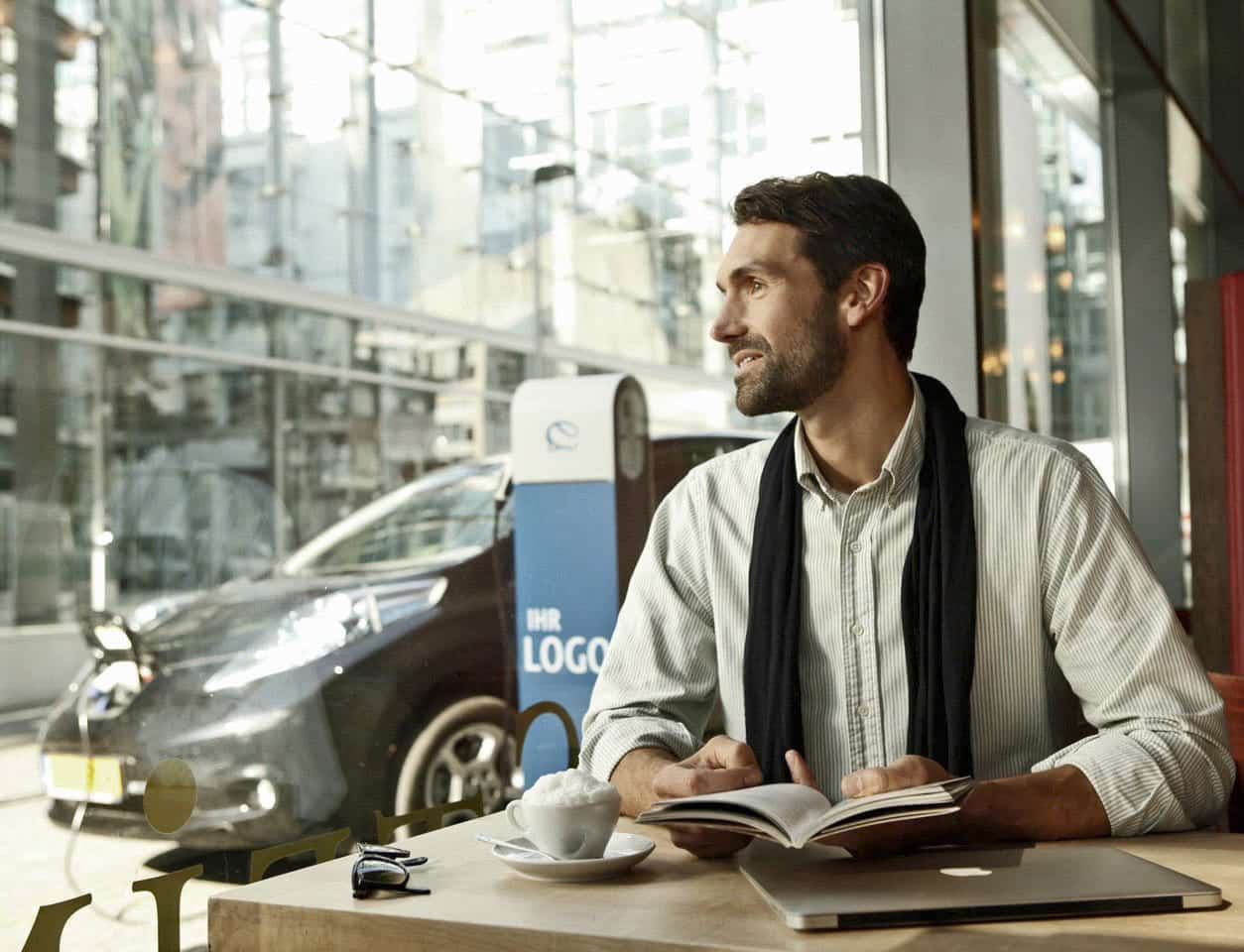 Ein Mann sitzt mit einem Notebook an einem Cafétisch und schaut aus dem Fenster. Er hat einen Bart und trägt einen Schal über seinem Hemd. Auf dem Tisch stehen eine Tasse Kaffee und eine Sonnenbrille. Draußen parkt ein Elektroauto an einer Ladestation. © Fotografie Tomas Rodriguez