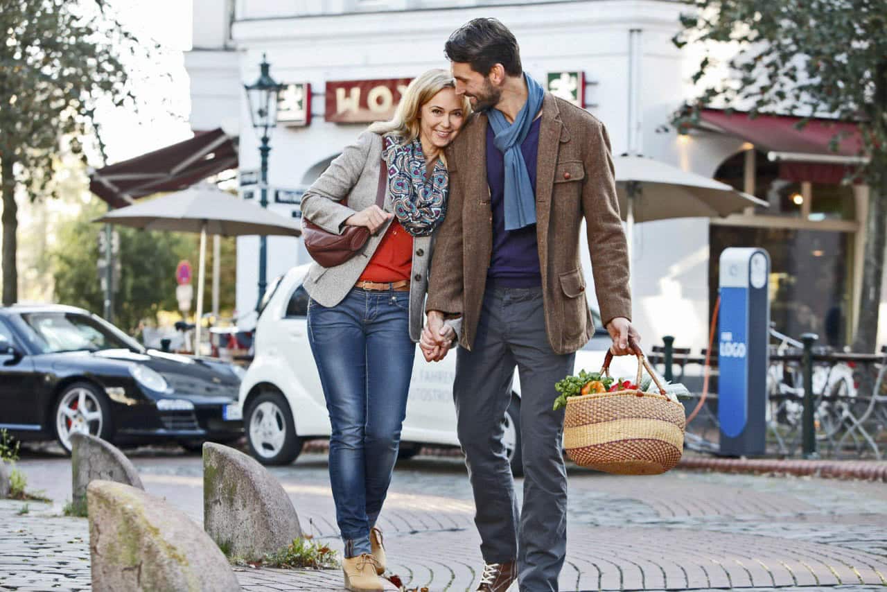 Ein Paar schlendert Hand in Hand durch eine Kopfsteinpflasterstraße, beide tragen legere Herbstkleidung. Der Mann trägt einen Weidenkorb voller Lebensmittel oder Obst und Gemüse. Bäume und geparkte Autos säumen die Straße, im Hintergrund sind ein Gebäude und Sitzgelegenheiten im Freien zu sehen. © Fotografie Tomas Rodriguez