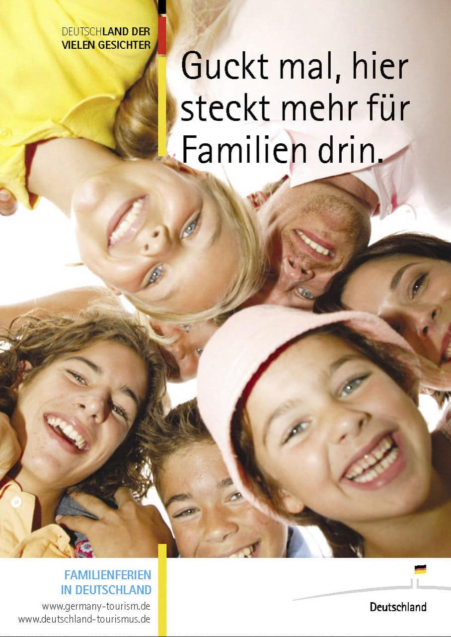 Eine Gruppe lächelnder Kinder und Erwachsener bildet einen Kreis und blickt in die Kamera. Der Text lautet: „Guckt mal, hier steckt mehr für Familien drin.“ Das Design wirbt für Familienurlaube in Deutschland und enthält einen Website-Link. © Fotografie Tomas Rodriguez