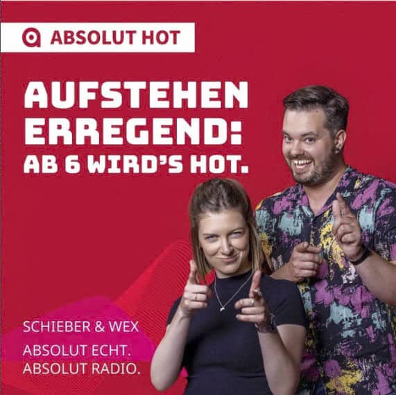 Zwei Personen stehen vor einem roten Hintergrund mit dem Schriftzug „Absolut Hot“. Sie gestikulieren, als würden sie auf den Betrachter zeigen. Der Text lautet: „Aufstehen Erregend: Ab 6 Wird’s Hot.“ Zusätzlicher Text: „Schieber & Wex Absolut Echt. Absolut Radio. © Fotografie Tomas Rodriguez