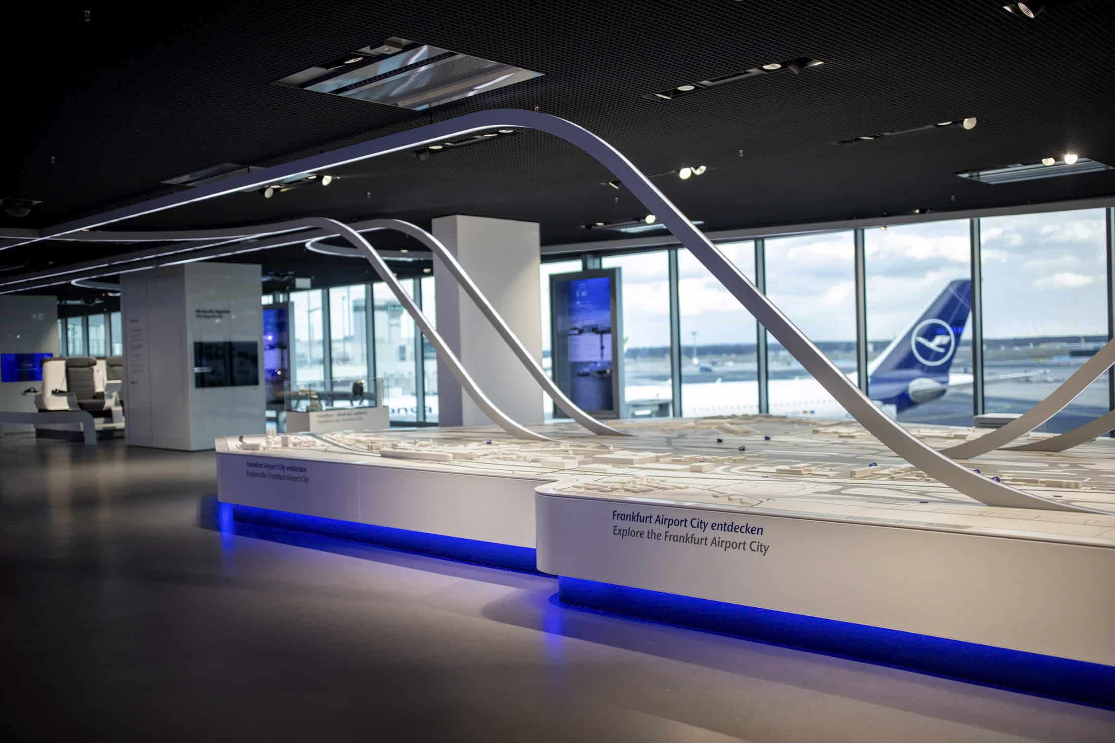 Modell einer Flughafenstadt in einem modernen Ausstellungsraum mit großen Fenstern, die den Blick auf ein Flugzeug und eine Landebahn draußen freigeben. Das Modell ist von unten blau beleuchtet und die Decke ist schwarz mit integrierter Beleuchtung. © Fotografie Tomas Rodriguez