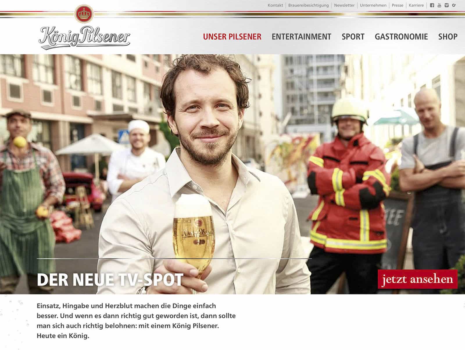 Ein Mann mit Bart und weißem Hemd hält ein Glas Bier in der Hand und lächelt. Hinter ihm stehen ein Feuerwehrmann, ein Bauarbeiter und ein Koch, alle lächelnd. Im Hintergrund ist eine Straßenszene zu sehen. Der Text wirbt für einen neuen TV-Spot für König Pilsener. © Fotografie Tomas Rodriguez