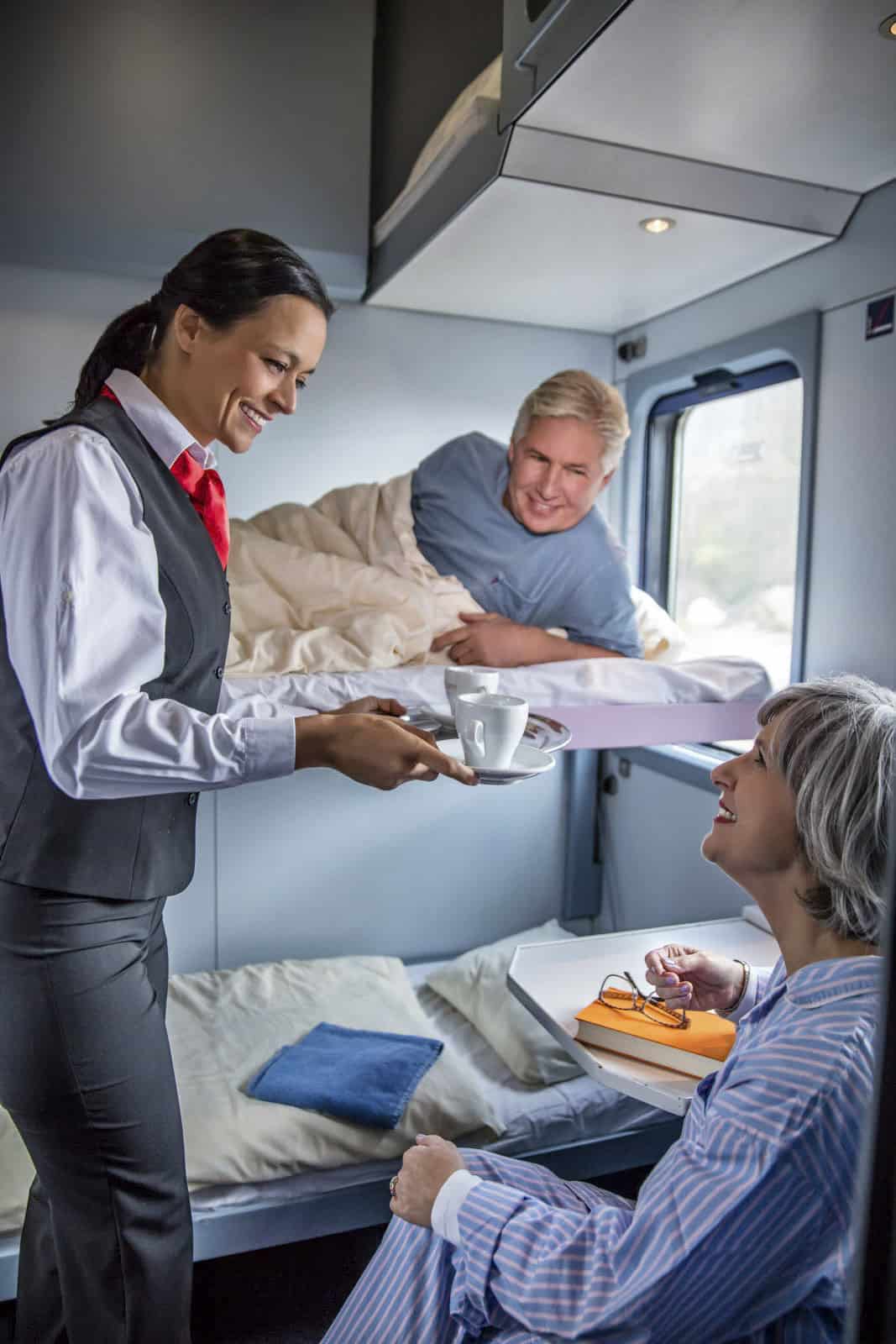 Ein Zugbegleiter serviert einer sitzenden Frau mit kurzen grauen Haaren, die ein Buch und eine Brille in der Hand hält, eine Tasse auf einem Tablett, während ein Mann mit blondem Haar auf dem oberen Etagenbett liegt, zusieht und lächelt. Die Kabine ist hell erleuchtet. © Fotografie Tomas Rodriguez