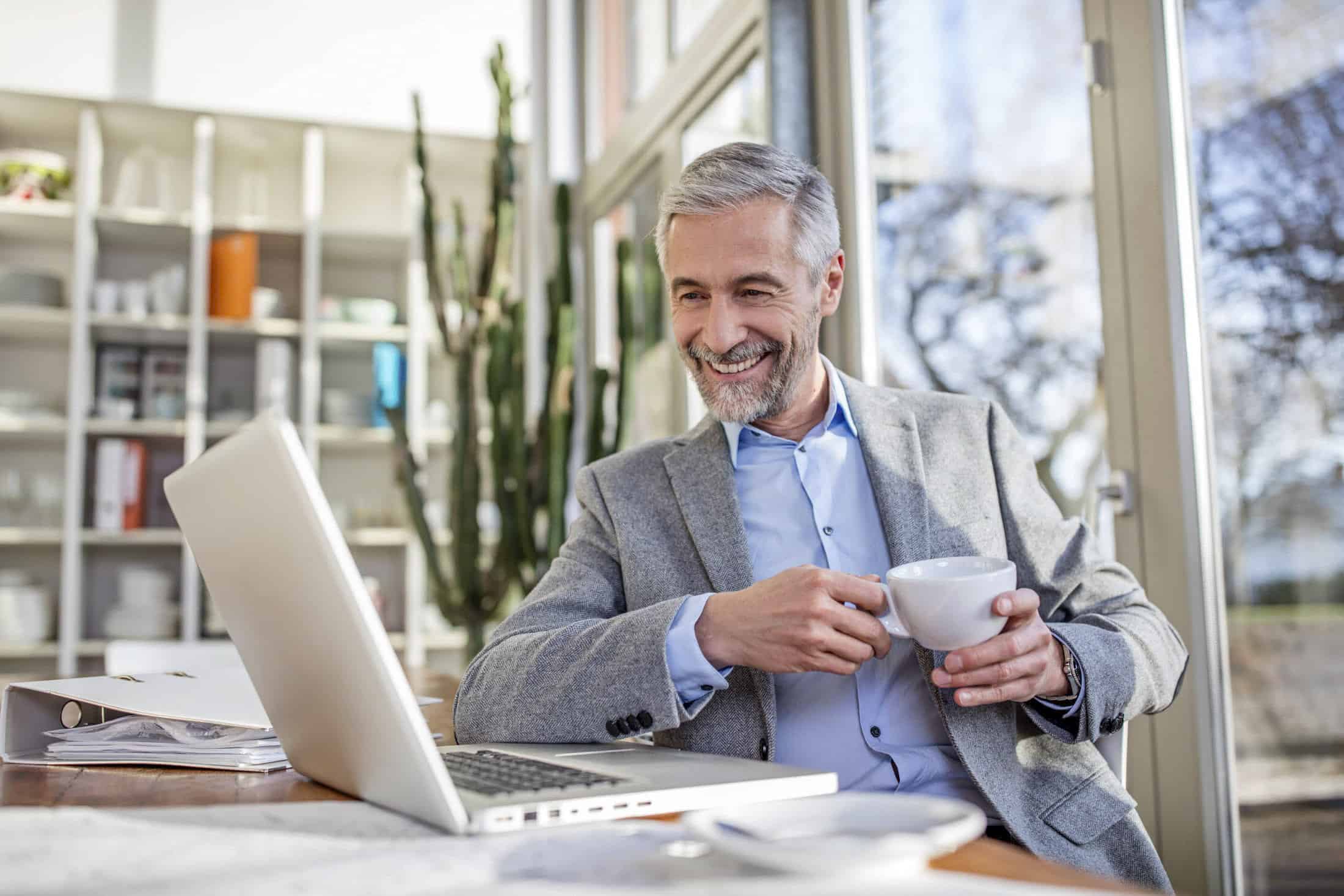 Ein lächelnder Mann mit grauem Haar und Bart sitzt an einem Tisch, hält eine Tasse Kaffee in der Hand und schaut auf einen Laptop. Er trägt ein graues Jackett. Der Raum hat große Fenster und Regale mit verschiedenen Gegenständen im Hintergrund. © Fotografie Tomas Rodriguez