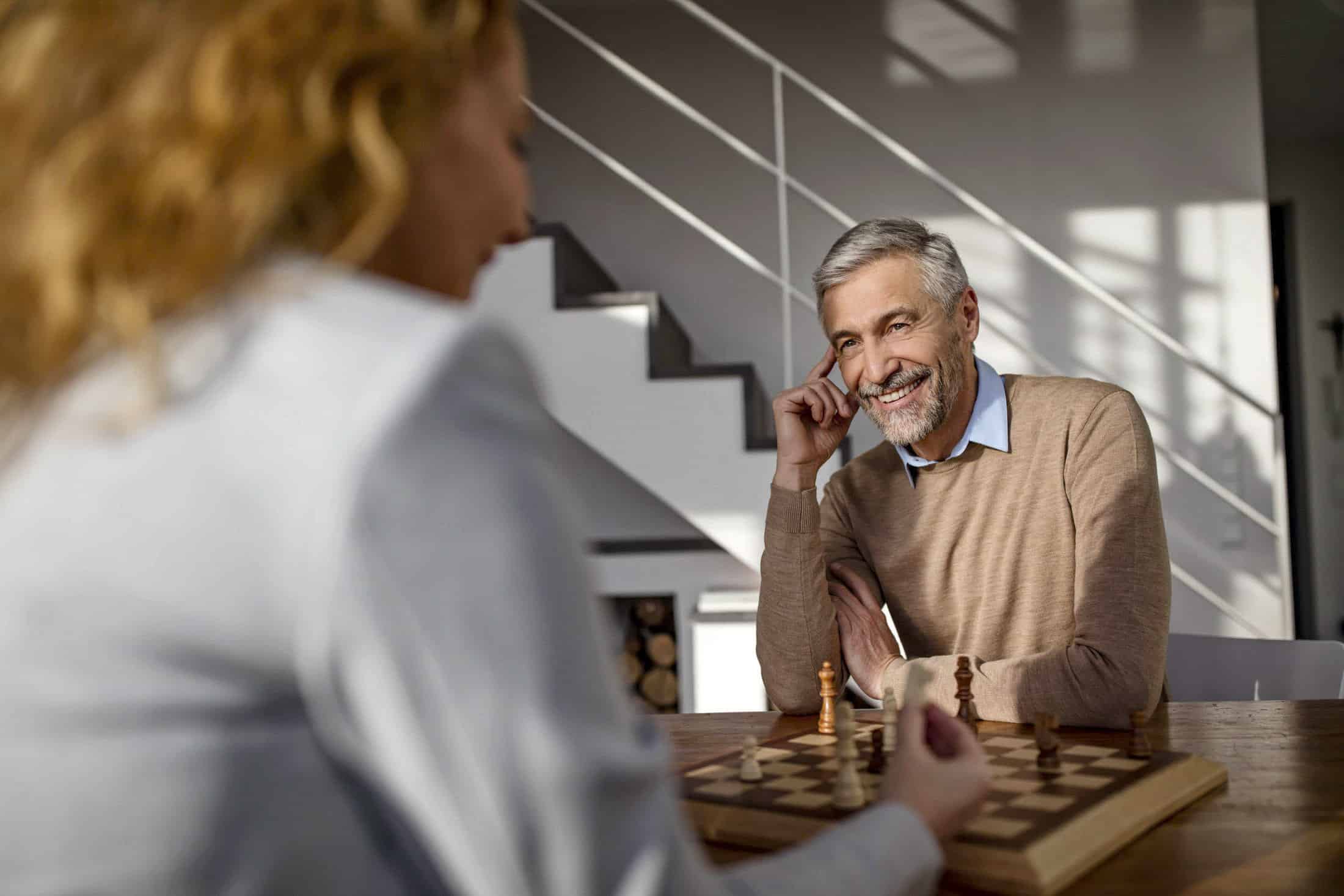 Ein Mann mit grauem Haar und Bart, der einen beigen Pullover trägt, sitzt an einem Tisch und spielt Schach. Er lächelt, während er Blickkontakt mit einer Frau im Vordergrund aufnimmt. Im Hintergrund sind eine Treppe und Regale zu sehen. © Fotografie Tomas Rodriguez