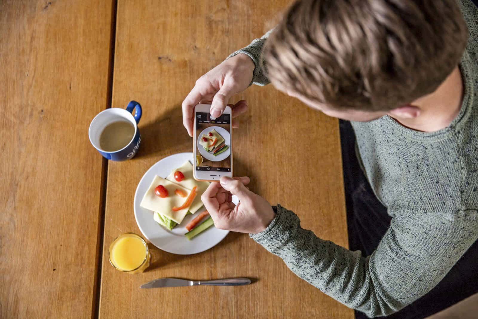 Eine Person im grünen Pullover fotografiert mit einem Smartphone einen Teller mit Käse, Kirschtomaten und geschnittenem Gemüse. In der Nähe liegen eine Tasse Kaffee, ein Glas Orangensaft und ein Messer auf einem Holztisch. © Fotografie Tomas Rodriguez