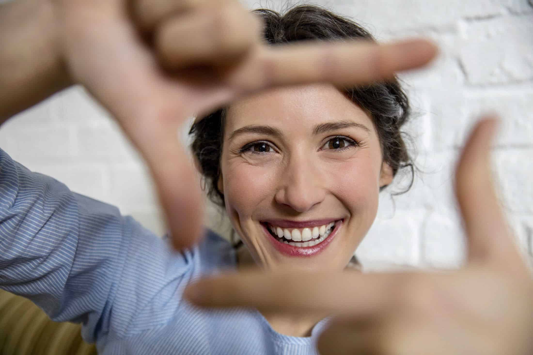 Eine Frau mit fröhlichem Gesichtsausdruck formt mit ihren Fingern ein Rechteck. Sie trägt ein hellblaues Hemd und steht vor einer weißen Backsteinwand. Ihre Finger bilden einen verspielten Rahmen um ihr lächelndes Gesicht. © Fotografie Tomas Rodriguez