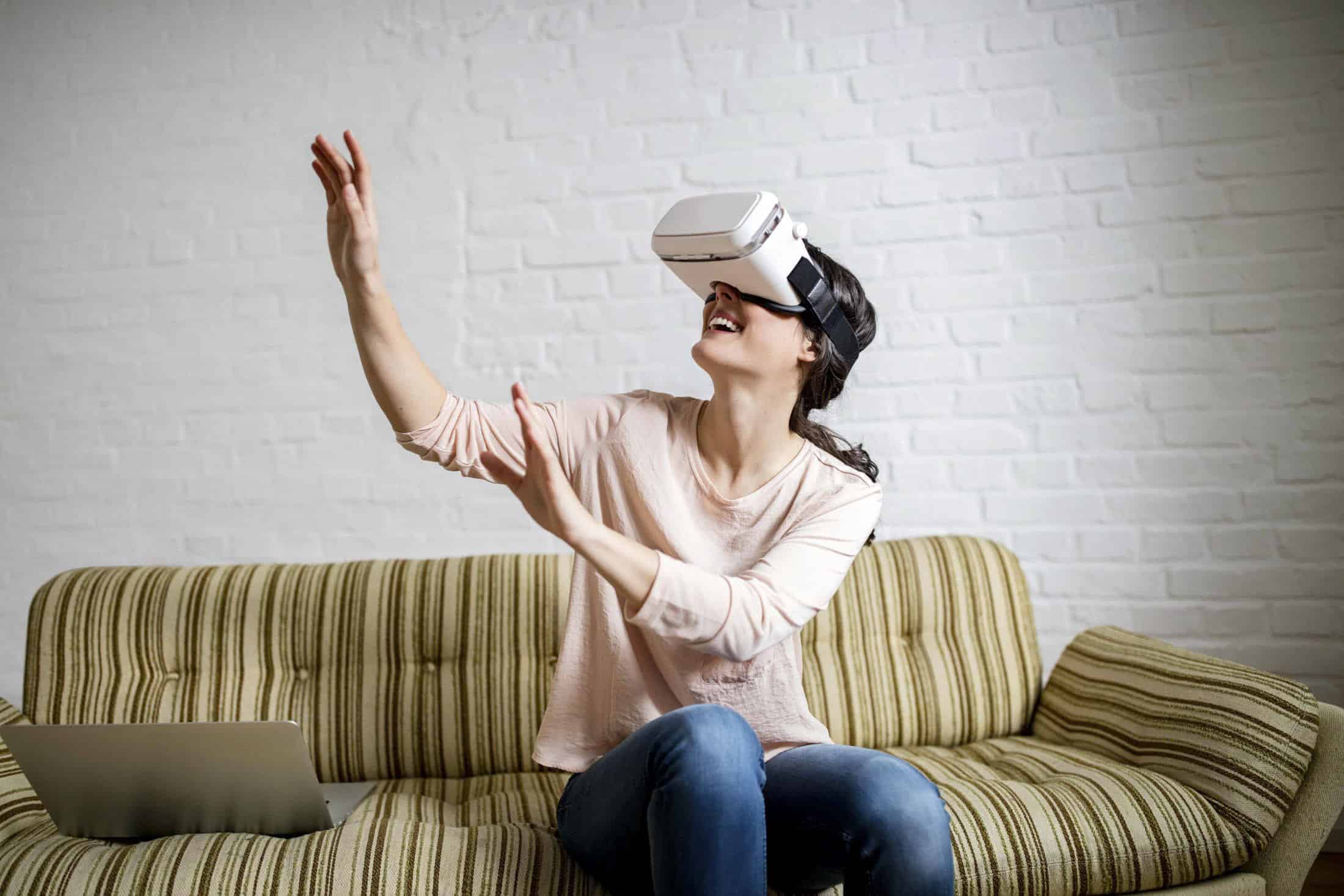 Frau sitzt auf einem gestreiften Sofa, trägt ein Virtual-Reality-Headset und streckt beide Hände aus, während sie lächelt. Neben ihr auf dem Sofa steht ein Laptop. Sie ist leger gekleidet und im Hintergrund ist eine weiße Backsteinwand. © Fotografie Tomas Rodriguez