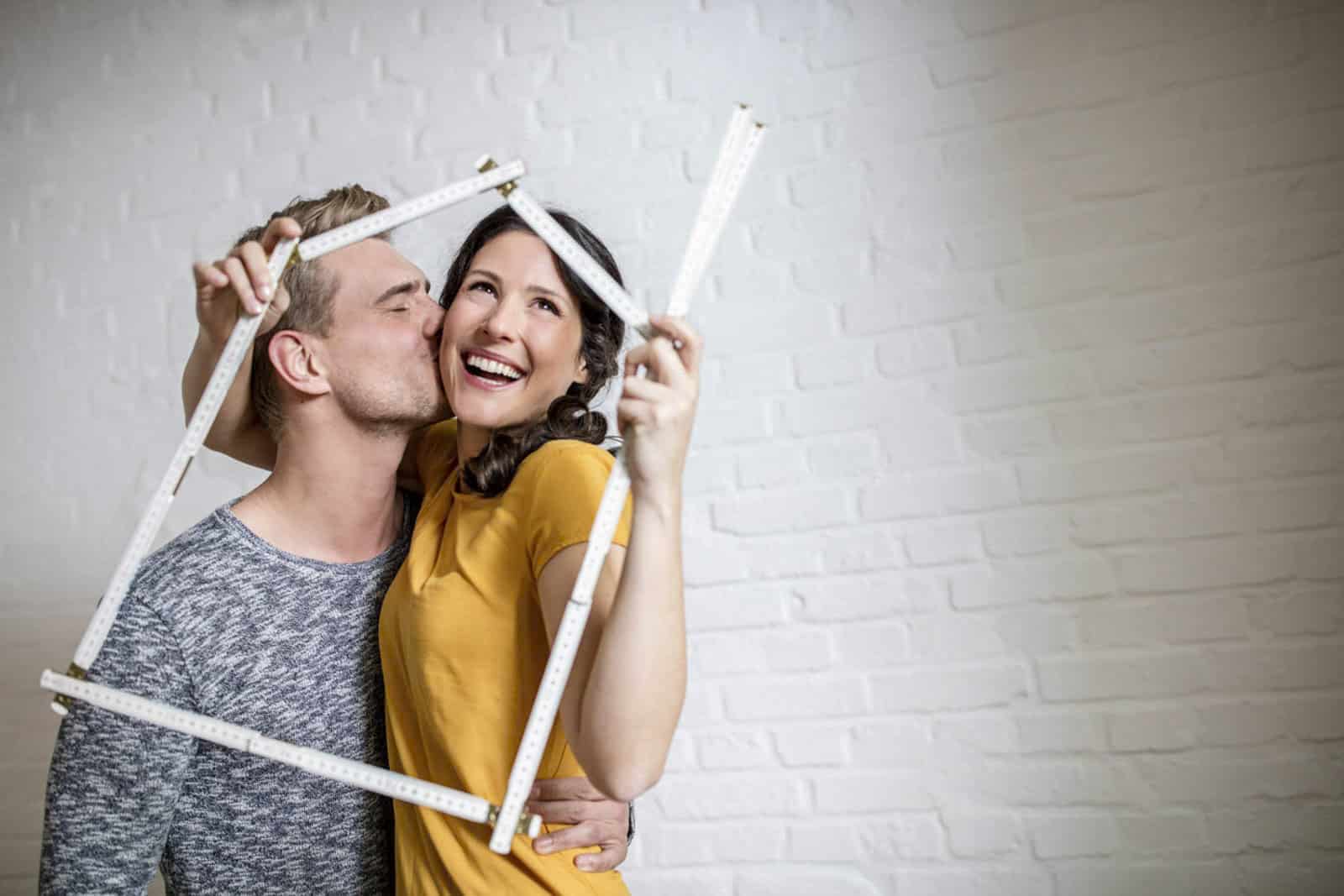 Ein lächelndes Paar posiert verspielt mit einem faltbaren Lineal, das einen quadratischen Rahmen bildet. Der Mann küsst die Frau auf die Wange, während sie vor einer weißen Backsteinwand stehen. Sie trägt ein gelbes Hemd und er ein grau gemustertes Hemd. © Fotografie Tomas Rodriguez