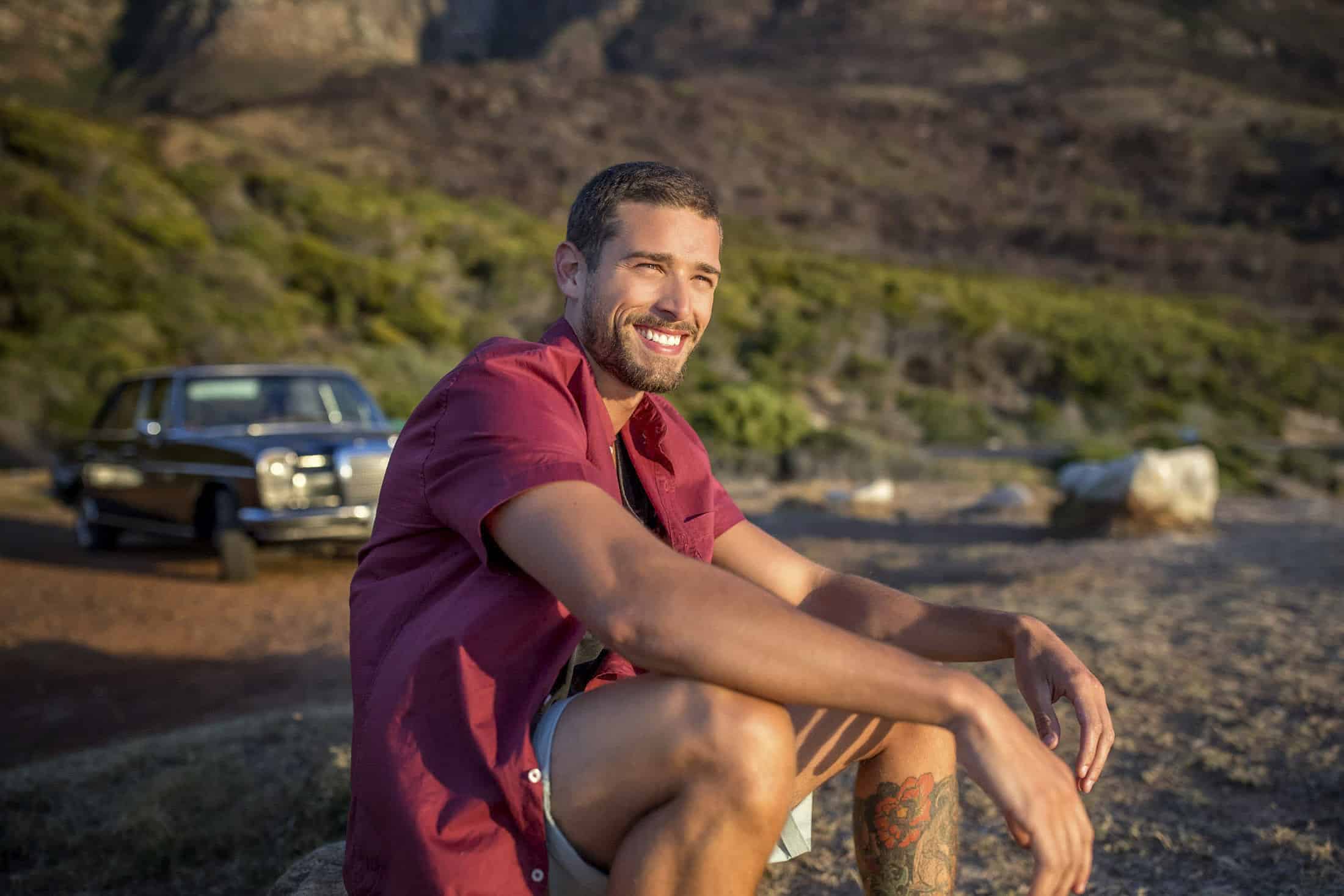Ein Mann mit kurzen dunklen Haaren und Bart sitzt lächelnd auf einem Felsen und trägt ein rotes Kurzarmhemd und Shorts. In der Nähe parkt an einem sonnigen Tag in einer natürlichen Landschaft mit Hügeln und Grünflächen ein Oldtimer. © Fotografie Tomas Rodriguez