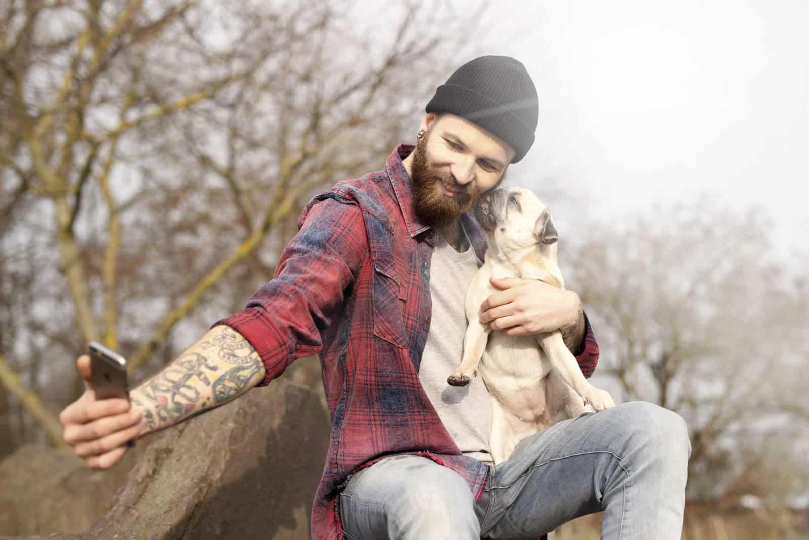 Ein bärtiger Mann mit Beanie und kariertem Hemd macht draußen ein Selfie, während er einen kleinen Mops hält. Die Szene spielt in einem Park mit kahlen Bäumen im Hintergrund, was auf einen kühlen Tag schließen lässt. © Fotografie Tomas Rodriguez
