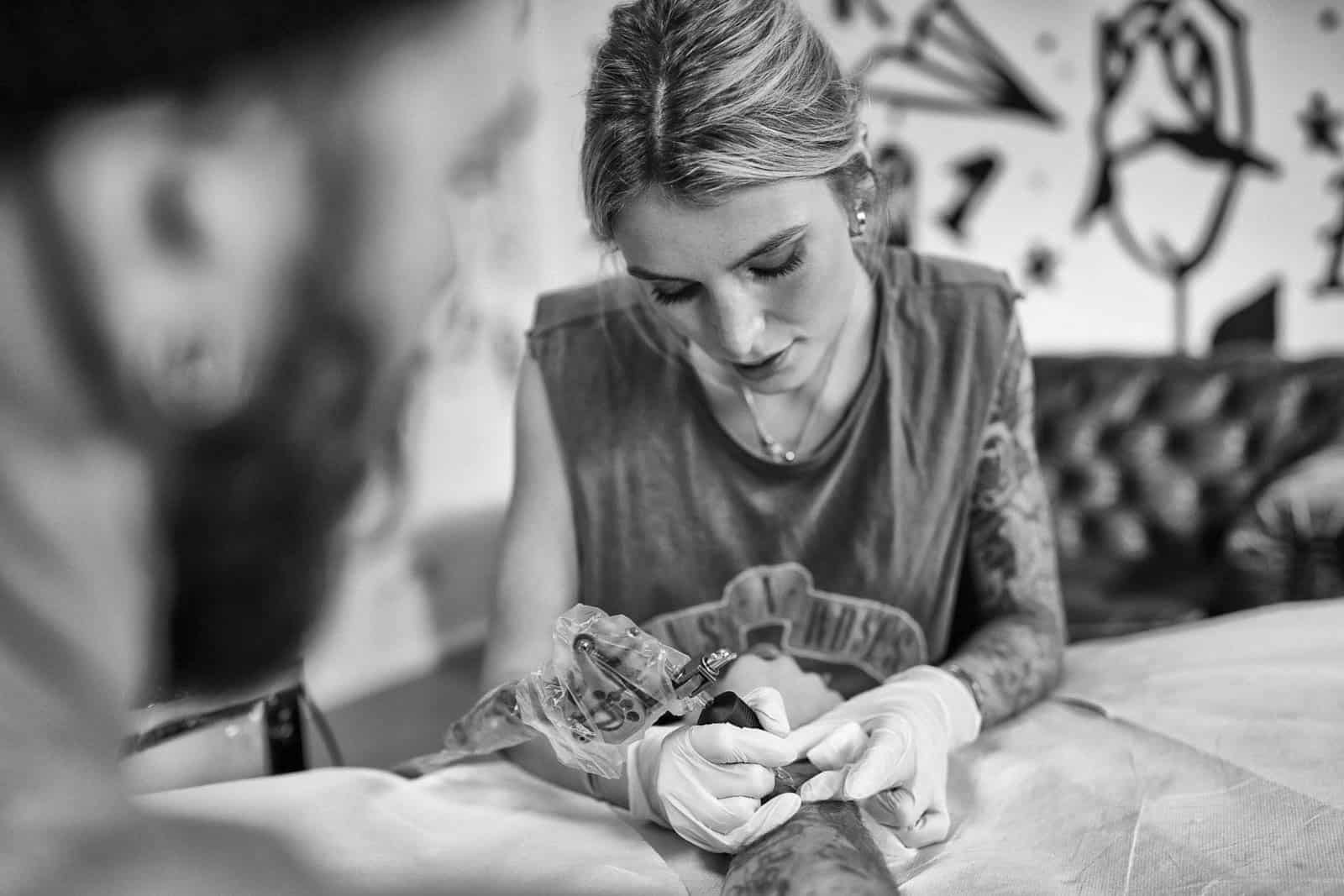 Eine Tätowiererin mit Tattoos auf den Armen ist darauf konzentriert, den Unterarm eines Kunden zu tätowieren. Sie trägt Handschuhe und legere Kleidung. Die Szene spielt in einem Tattoostudio mit verschiedenen Kunstwerken an den Wänden. Das Bild ist in Schwarzweiß. © Fotografie Tomas Rodriguez