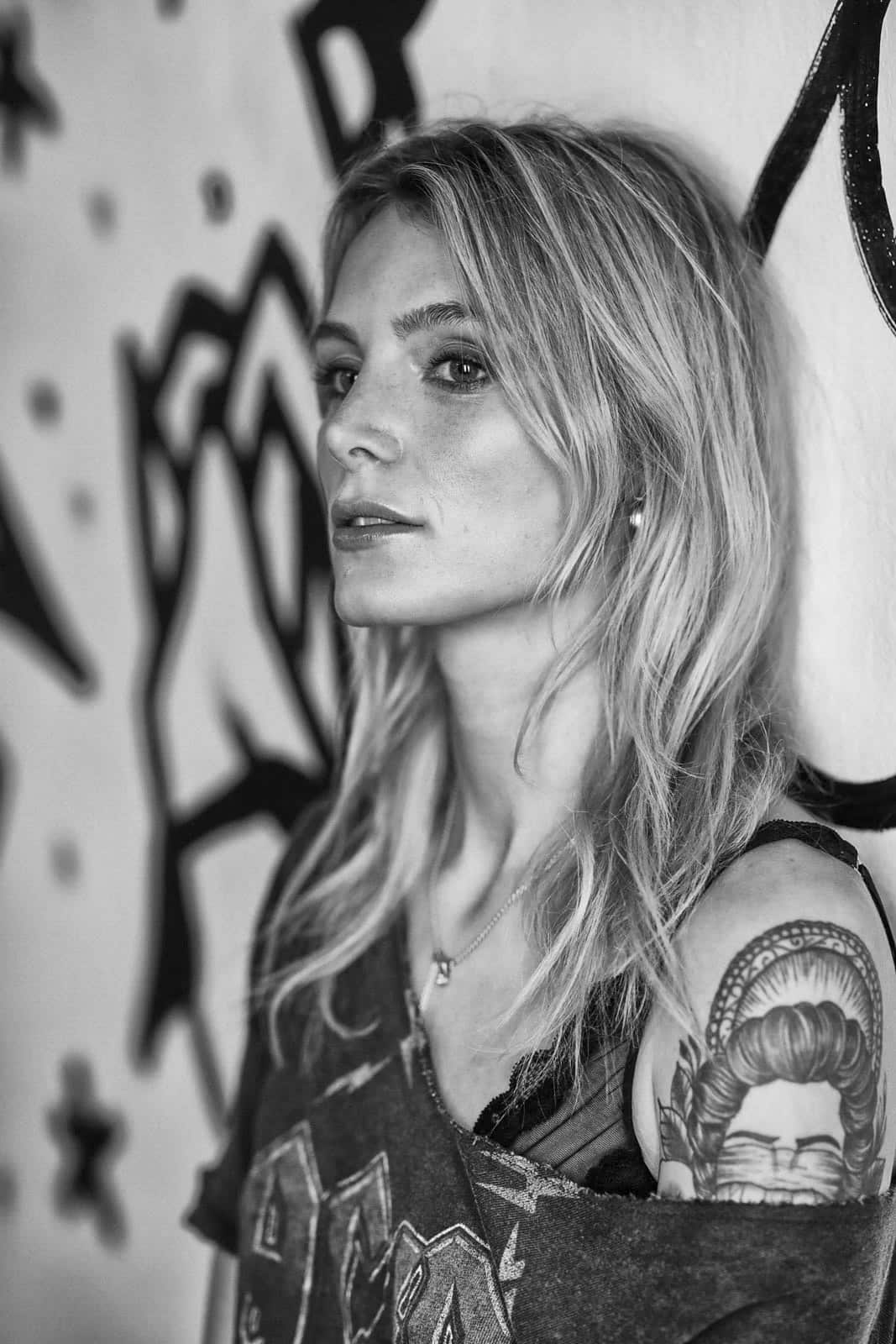 Schwarzweißbild einer Frau mit langen, welligen Haaren, die an einer Wand mit Graffiti lehnt. Sie hat ein sichtbares Tattoo auf dem Arm und trägt ein abgenutztes Tanktop. Ihr Gesichtsausdruck ist nachdenklich. © Fotografie Tomas Rodriguez