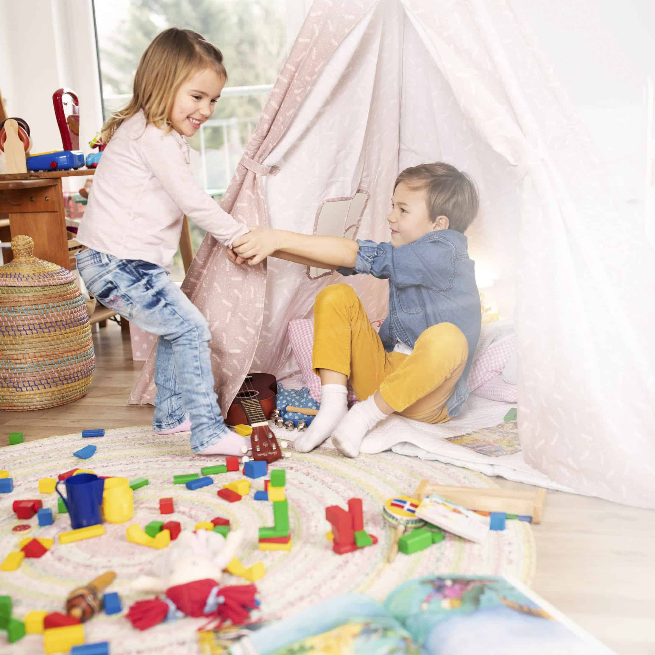 Zwei Kinder spielen in einem bunten Raum. Eines sitzt in einem Stoffspielzelt, das andere steht draußen und hält die Zeltklappe fest. Auf dem Boden liegen verstreut Spielsachen und Bauklötze, im Hintergrund ist ein geflochtener Korb zu sehen. © Fotografie Tomas Rodriguez