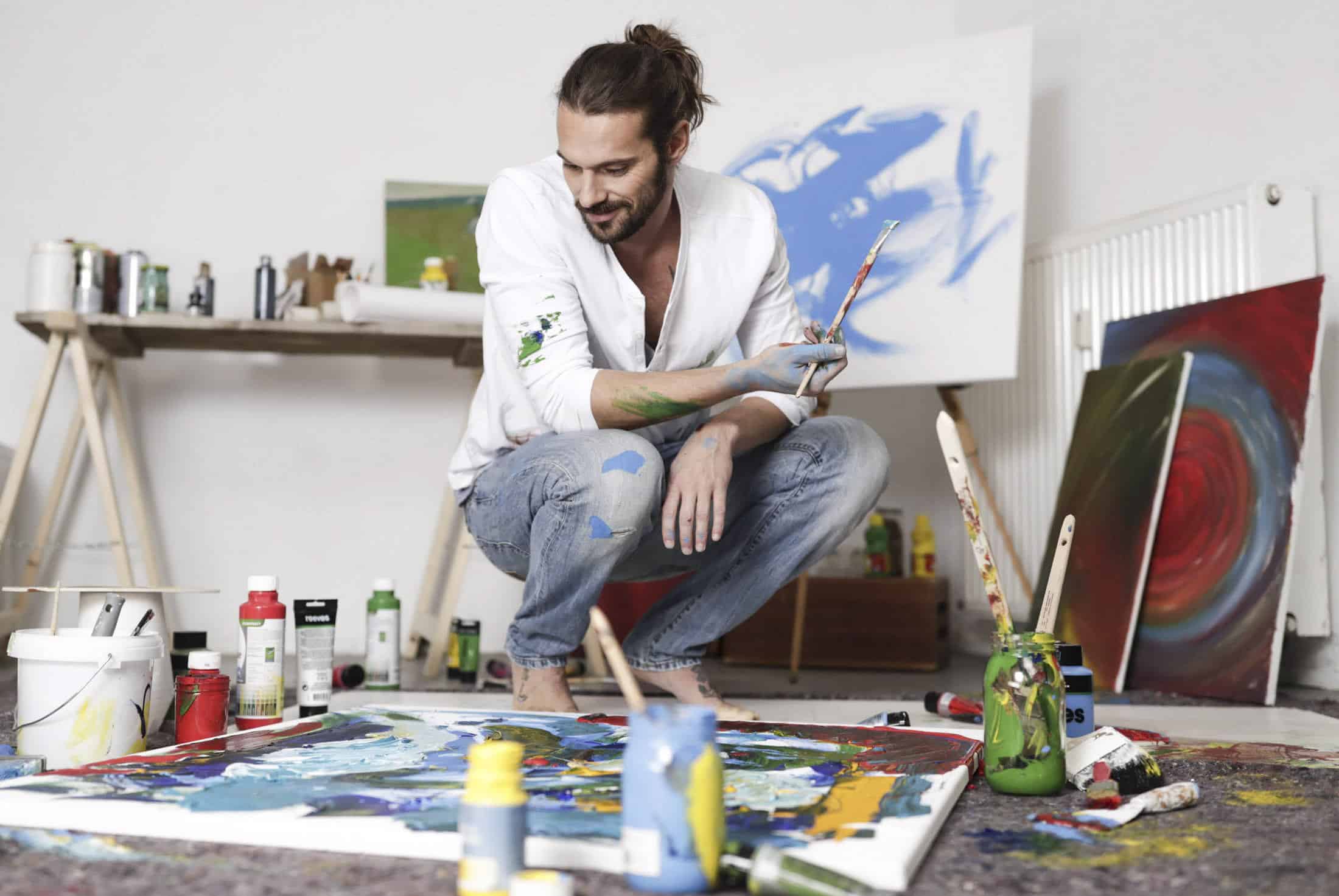 Ein Mann mit Dutt und weißem Hemd hockt auf dem Boden eines Ateliers und hält einen Pinsel in der Hand. Er ist umgeben von bunten Farben und einer großen Leinwand mit abstrakter Kunst. Im Hintergrund sind Leinwände und Malutensilien zu sehen. © Fotografie Tomas Rodriguez