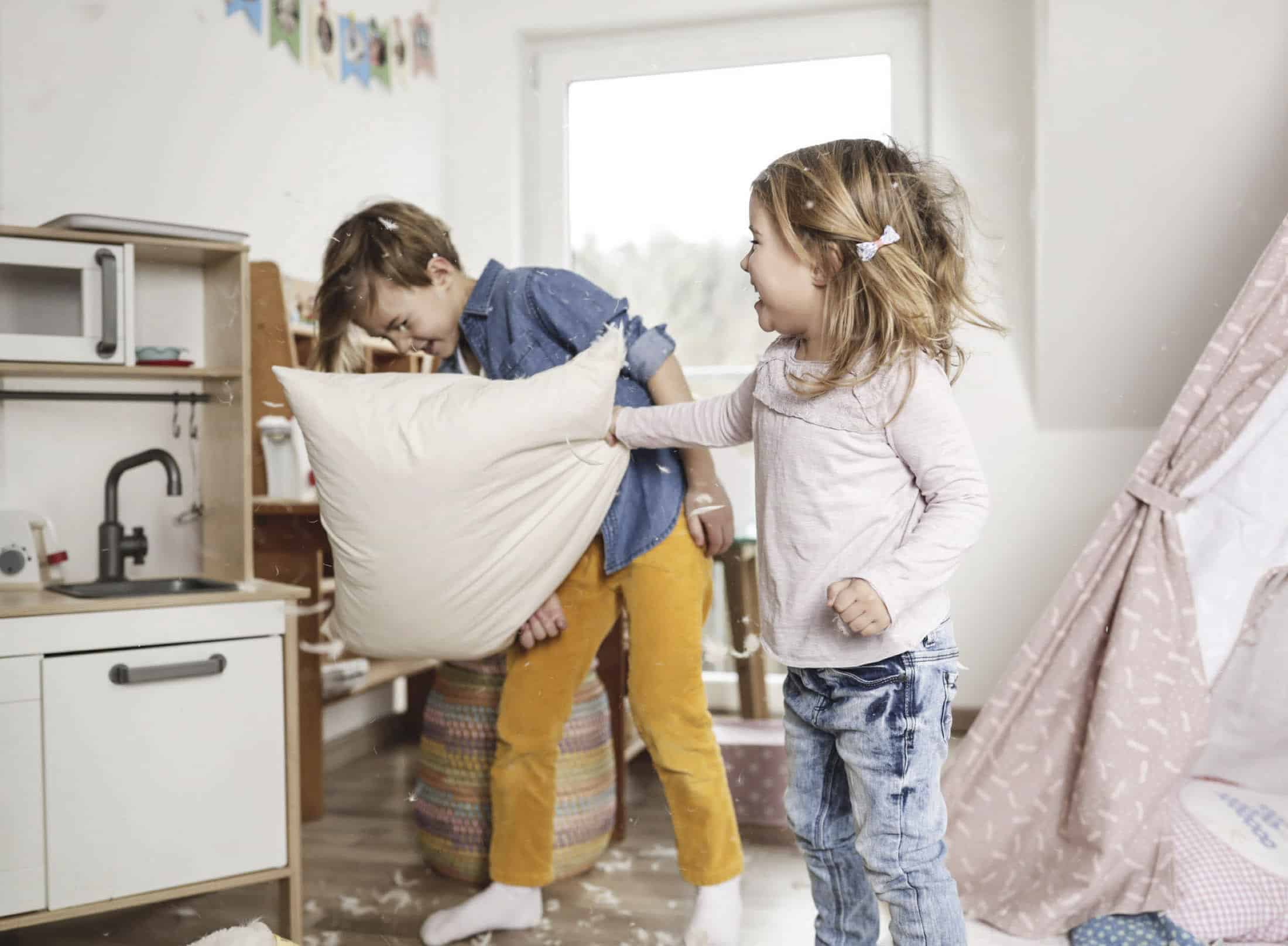 Zwei Kinder liefern sich in einem gemütlichen Zimmer spielerisch eine Kissenschlacht. Ein Junge in Jeanshemd und senffarbener Hose blickt abgewandt, während ein Mädchen in hellrosa Hemd und Jeans fröhlich ein Kissen schwingt, das von verstreuten Federn umgeben ist. © Fotografie Tomas Rodriguez