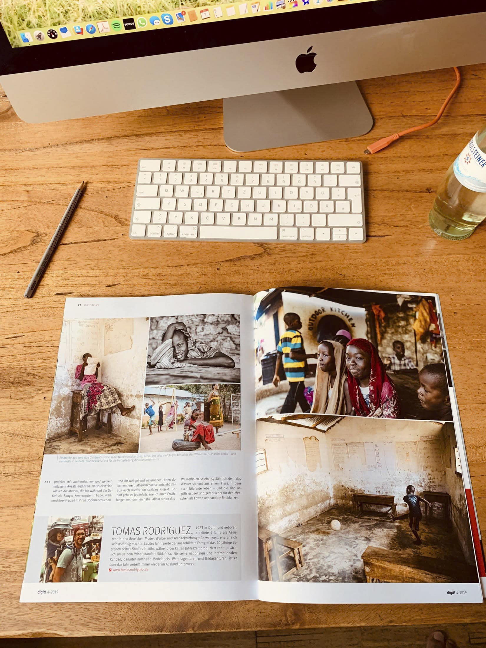 Eine aufgeschlagene Zeitschrift auf einem Holzschreibtisch unter einem iMac. Die linke Seite zeigt mehrere Fotos und Text über „Tomas Rodriguez“, während die rechte Seite Bilder von Menschen in lebhaften Umgebungen zeigt. Daneben liegen eine Tastatur, ein Bleistift und eine Getränkeflasche. © Fotografie Tomas Rodriguez