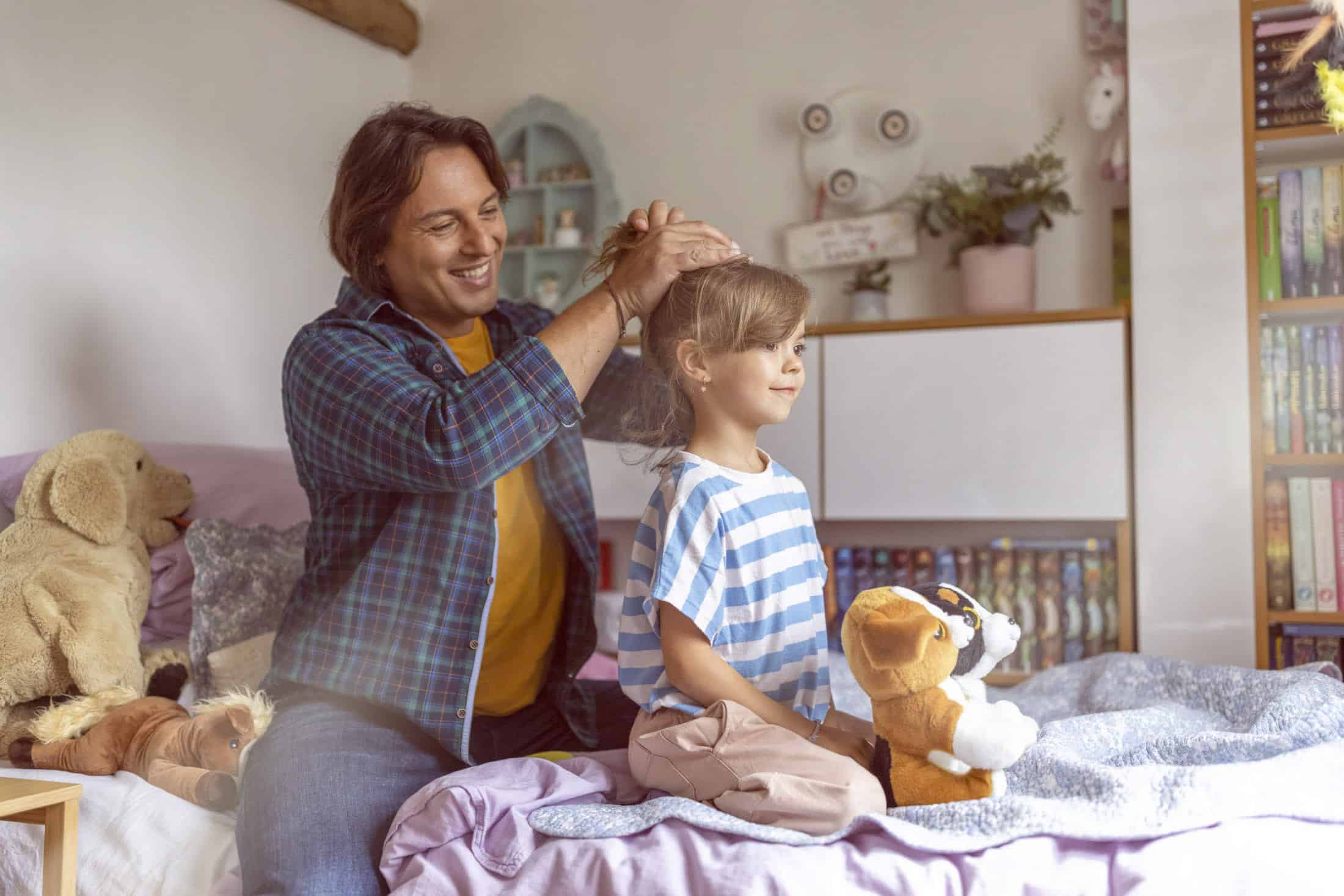 Ein Mann frisiert fröhlich einem kleinen Kind in einem gemütlichen Schlafzimmer voller Stofftiere und Bücher. Das Kind sitzt geduldig auf dem Bett und sanftes Sonnenlicht erhellt die Szene und schafft eine warme und freundliche Atmosphäre. © Fotografie Tomas Rodriguez
