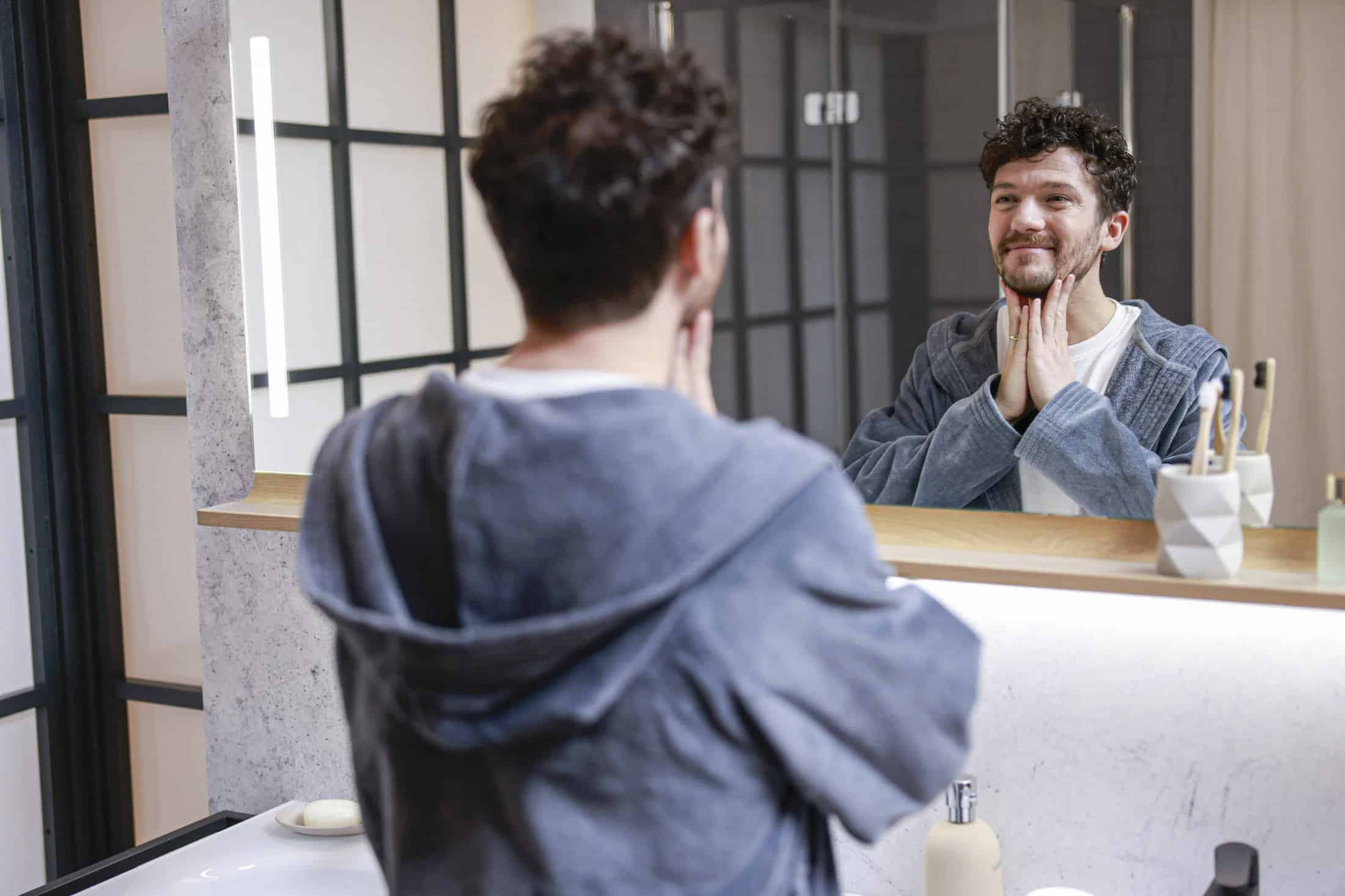 Ein Mann mit lockigem Haar betrachtet sich im Badezimmerspiegel. Er trägt einen grauen Bademantel. Er berührt sein Gesicht mit beiden Händen und lächelt leicht. Auf der Ablagefläche stehen ein Zahnbürstenhalter und eine Flasche. © Fotografie Tomas Rodriguez