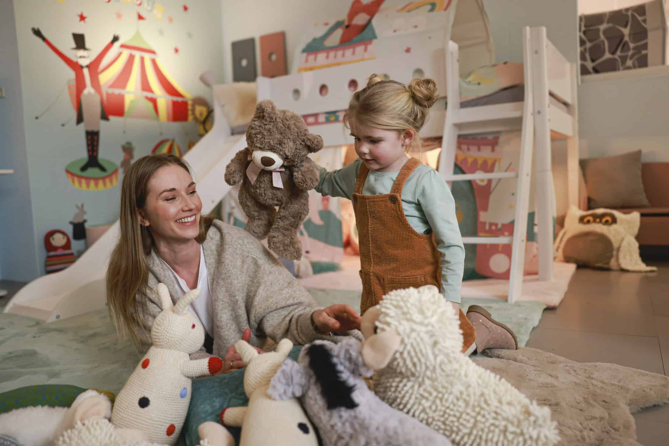 Frau und junges Mädchen spielen auf einem weichen Teppich mit Stofftieren in einem farbenfrohen Zimmer mit Zirkusthema. Die Frau lächelt und das Mädchen hält einen Teddybären. Im Hintergrund ist ein Etagenbett mit verspieltem Dekor zu sehen. © Fotografie Tomas Rodriguez