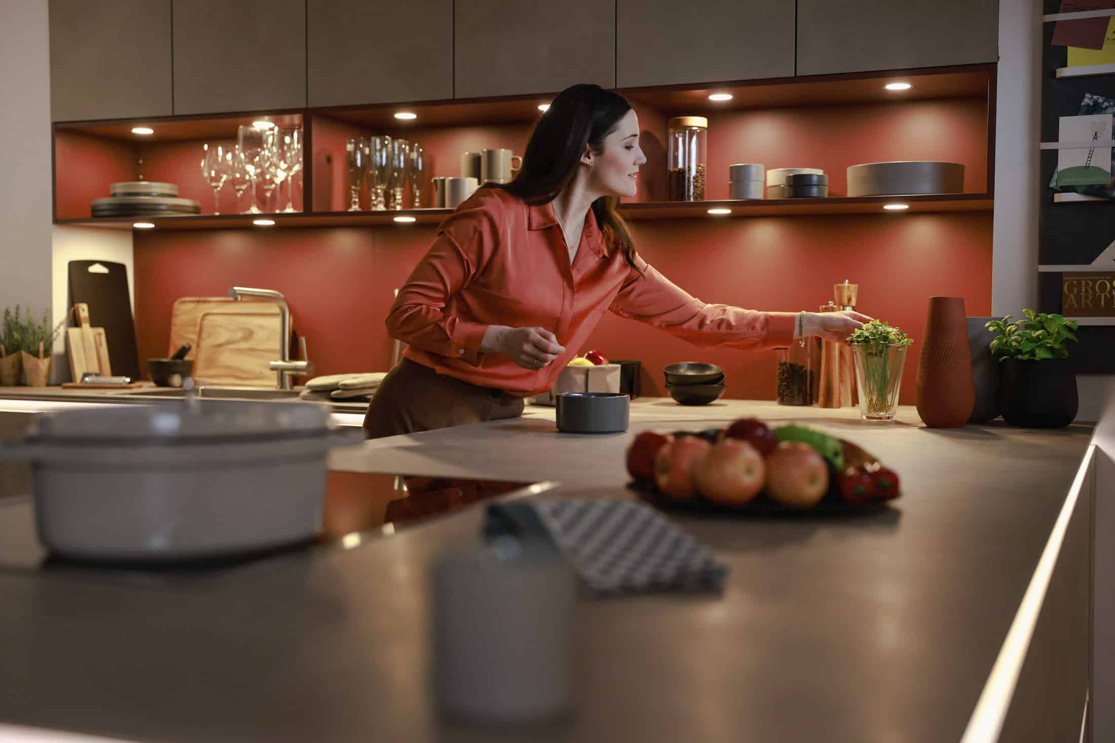 Eine Frau in einer roten Bluse ordnet Gegenstände auf einem Küchenregal. Die moderne Küche verfügt über rote Akzente, Regale mit Glaswaren und eine Arbeitsplatte mit einer Obstschale und einem gemusterten Tuch. Warmes Licht schafft eine gemütliche Atmosphäre. © Fotografie Tomas Rodriguez